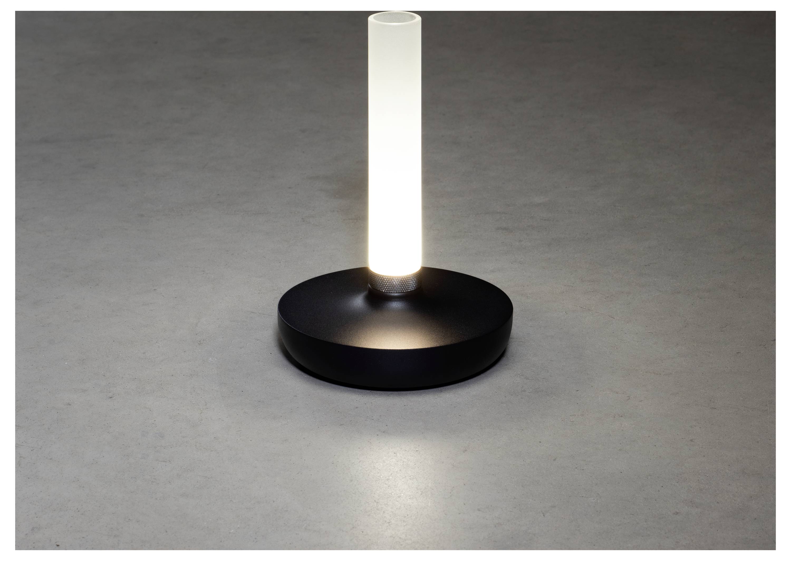 A modern, cylindrical table lamp emits soft white light from a frosted glass column, set on a simple, black circular base, against a gray surface.
