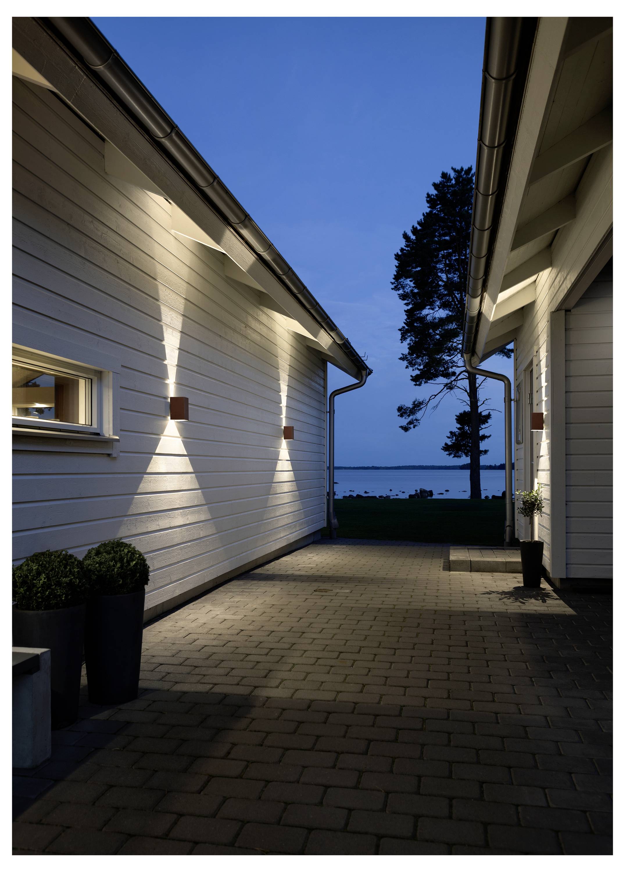 A narrow alley between two modern white houses, with wall lights casting beams on the facade, leading to a view of a lake at dusk.