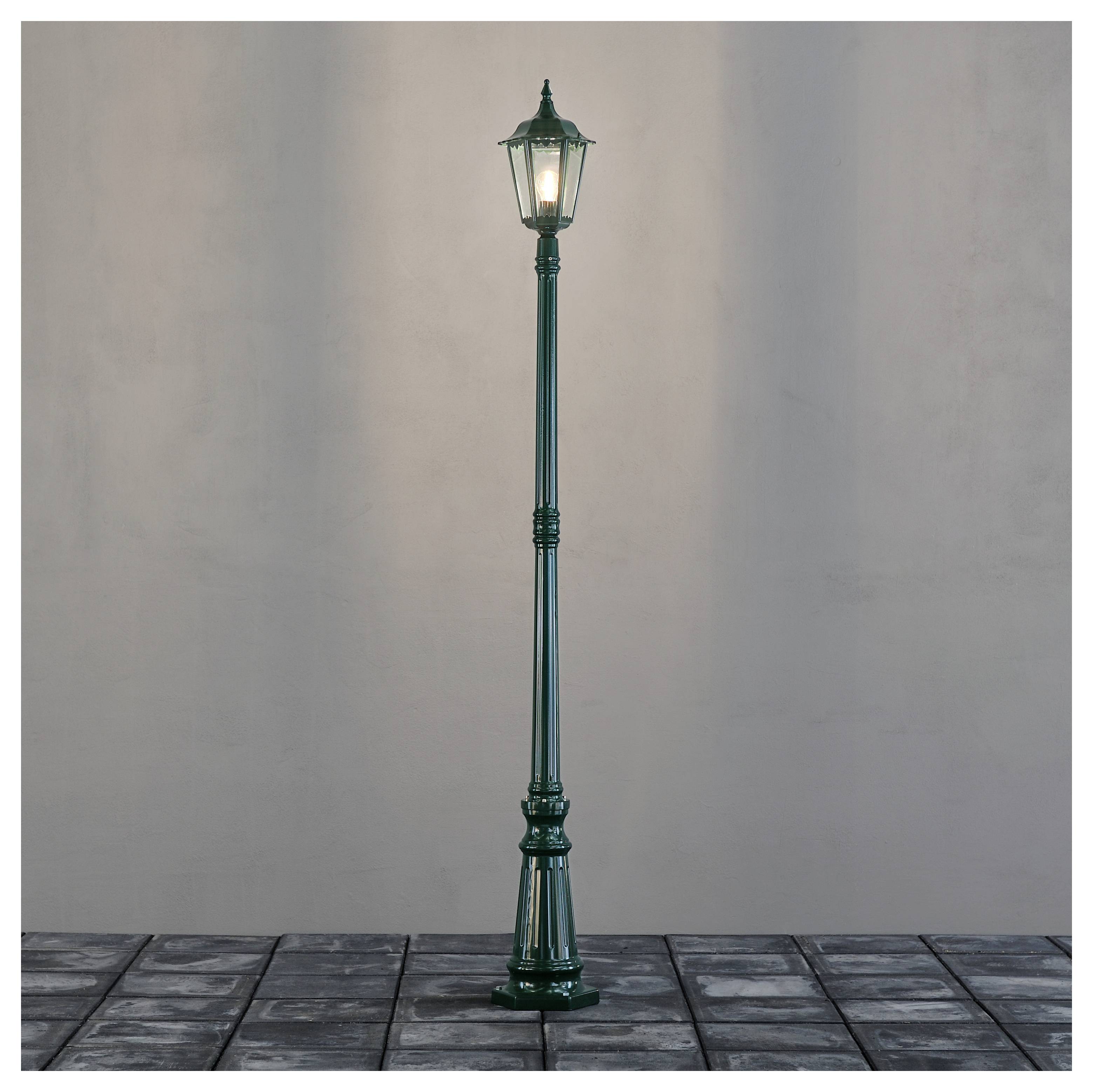 A single vintage-style street lamp with a glowing light, standing on a cobblestone path against a plain background.