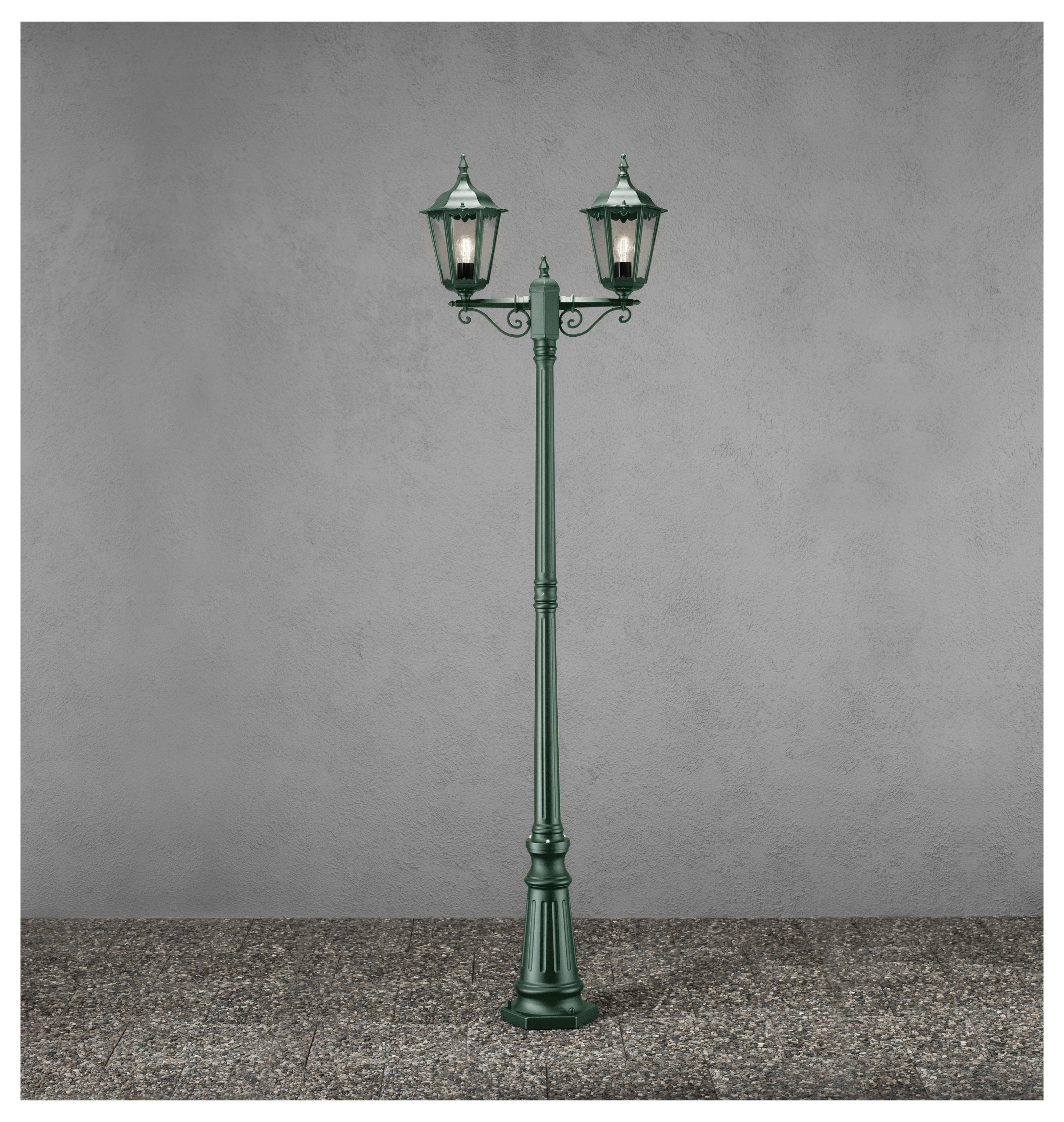 A classic green street lamp post with two illuminated lanterns stands on a cobblestone surface against a plain gray background.