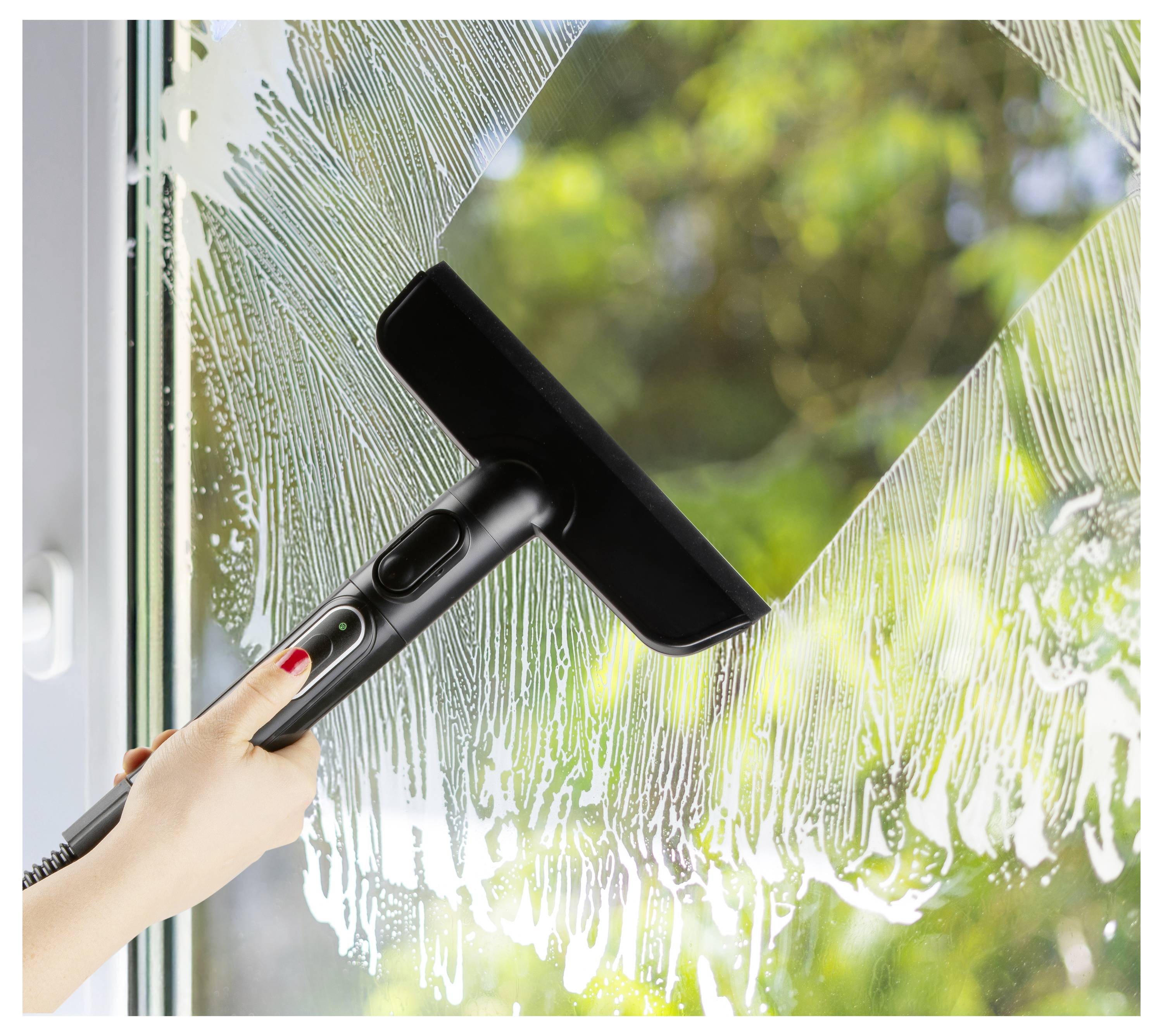 A hand using a black squeegee to clean soapy water from a window, revealing a clear view of green foliage outside.