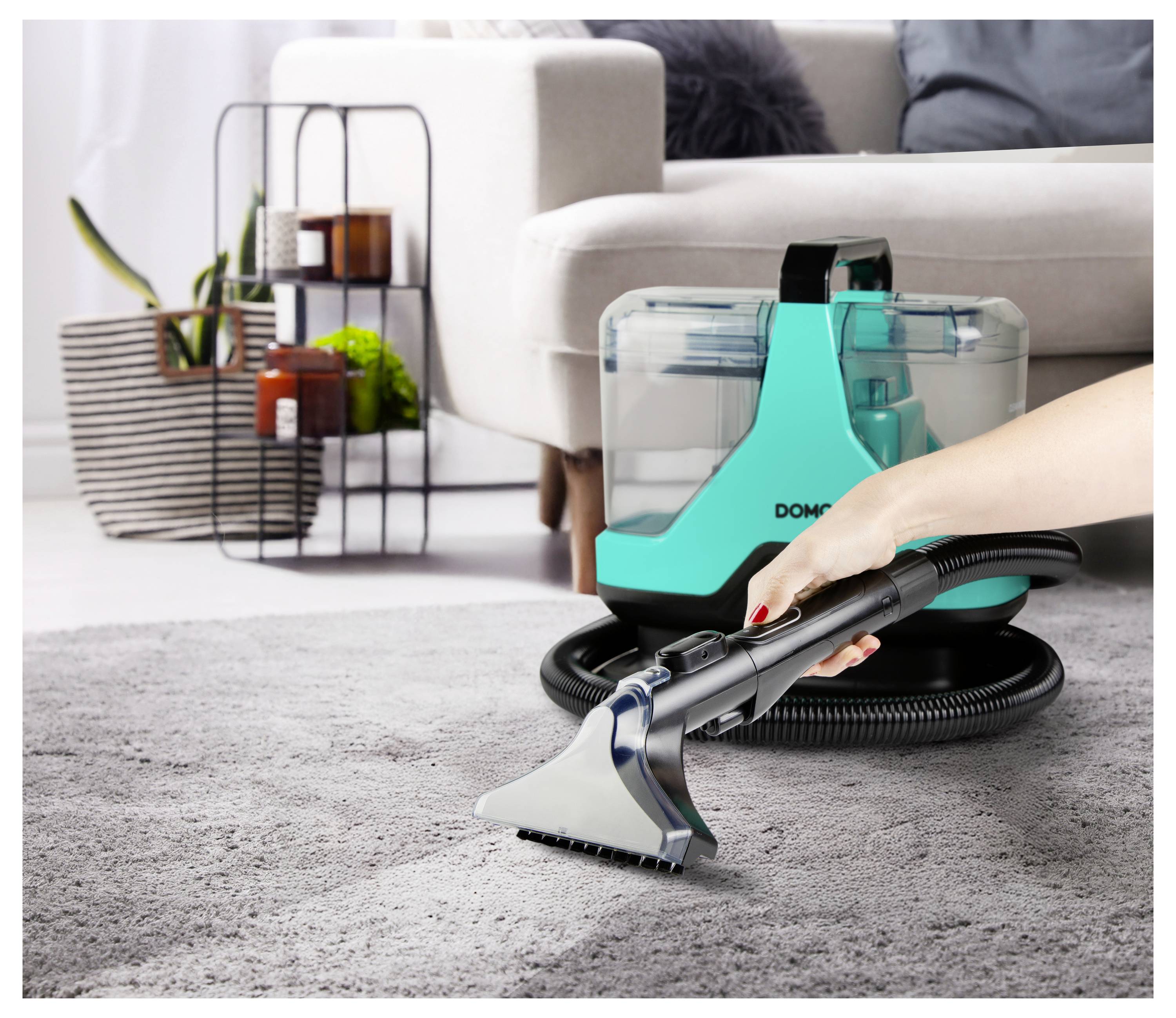 A person uses a portable carpet cleaner on a gray rug in a modern living room. The device features a transparent water tank.