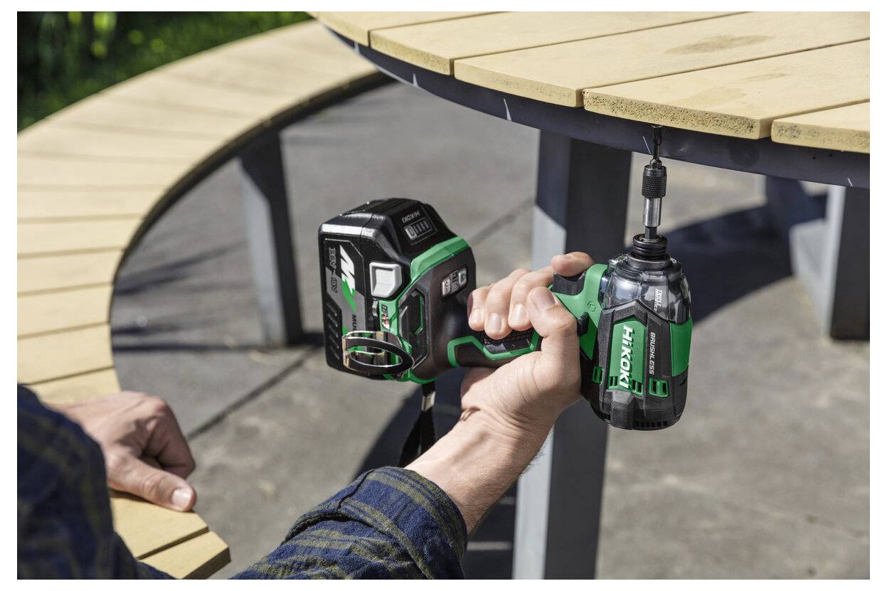 A person holds a green and black cordless drill near a round wooden table outdoors, demonstrating the tool in use.