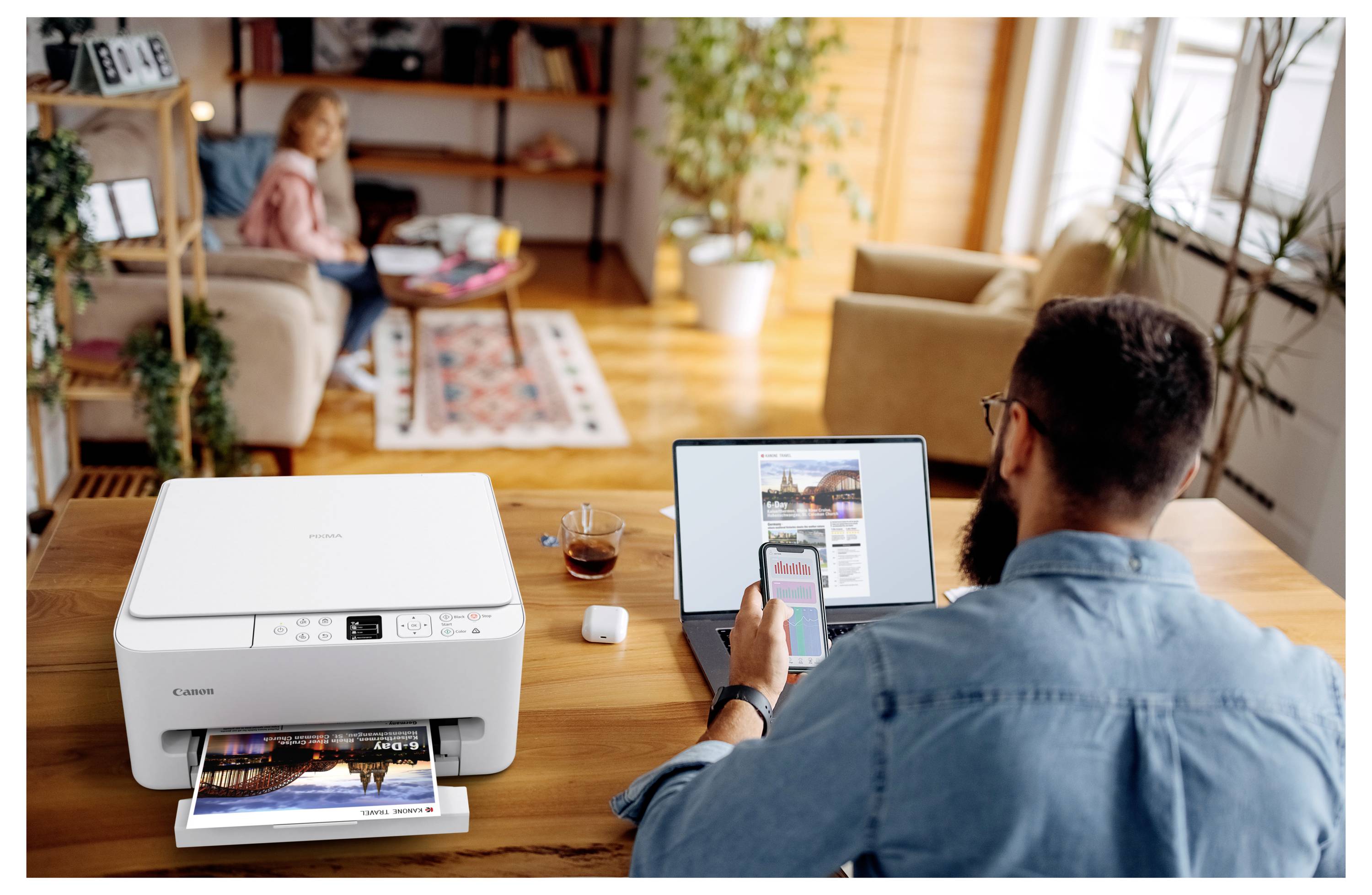 A man sits at a table using a smartphone and laptop to print a document on a nearby printer; a woman relaxes on a couch in the background.