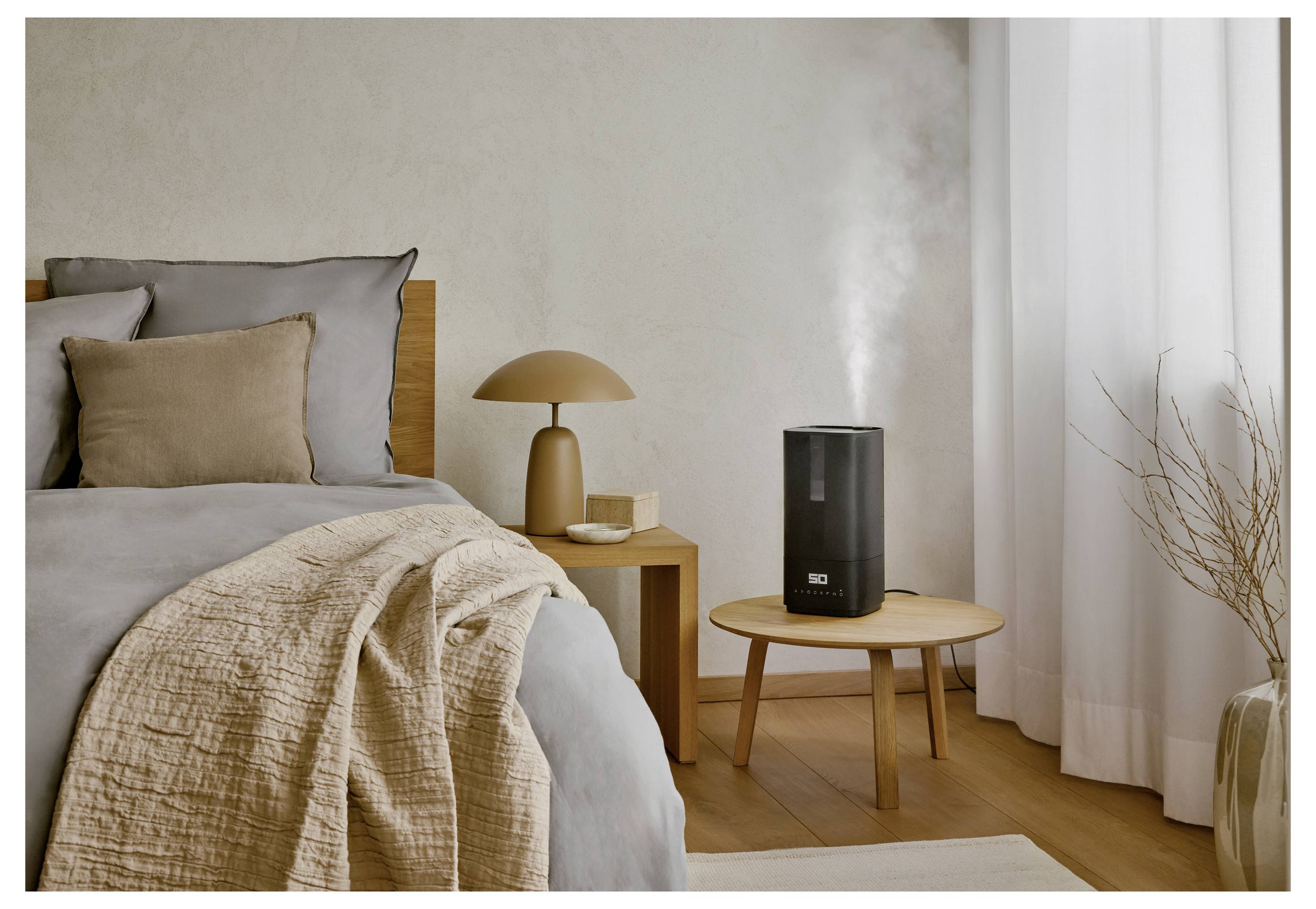 A modern bedroom features a bed with beige and gray bedding. A small wooden table holds a black humidifier emitting mist and a lamp.