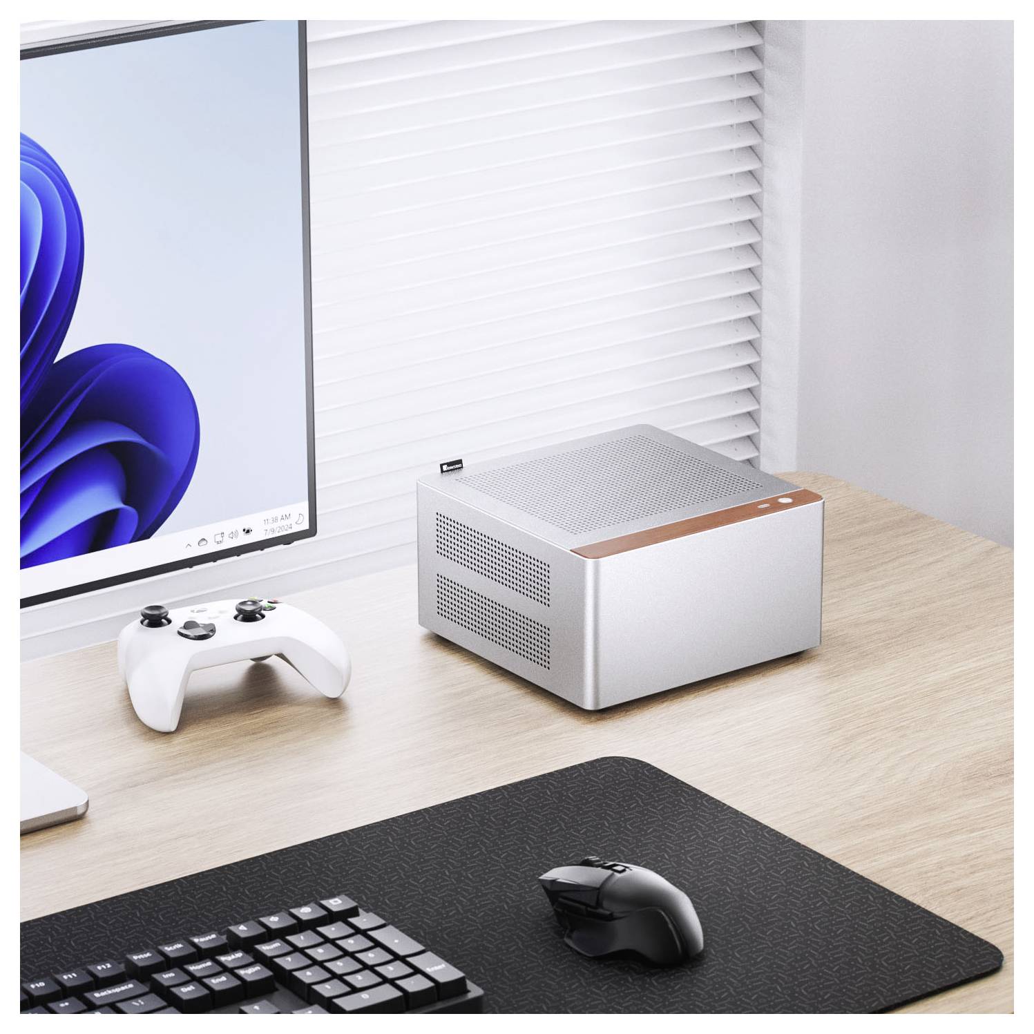 A compact desktop computer on a wooden desk, next to a keyboard and game controller, with a monitor displaying a blue swirl pattern.