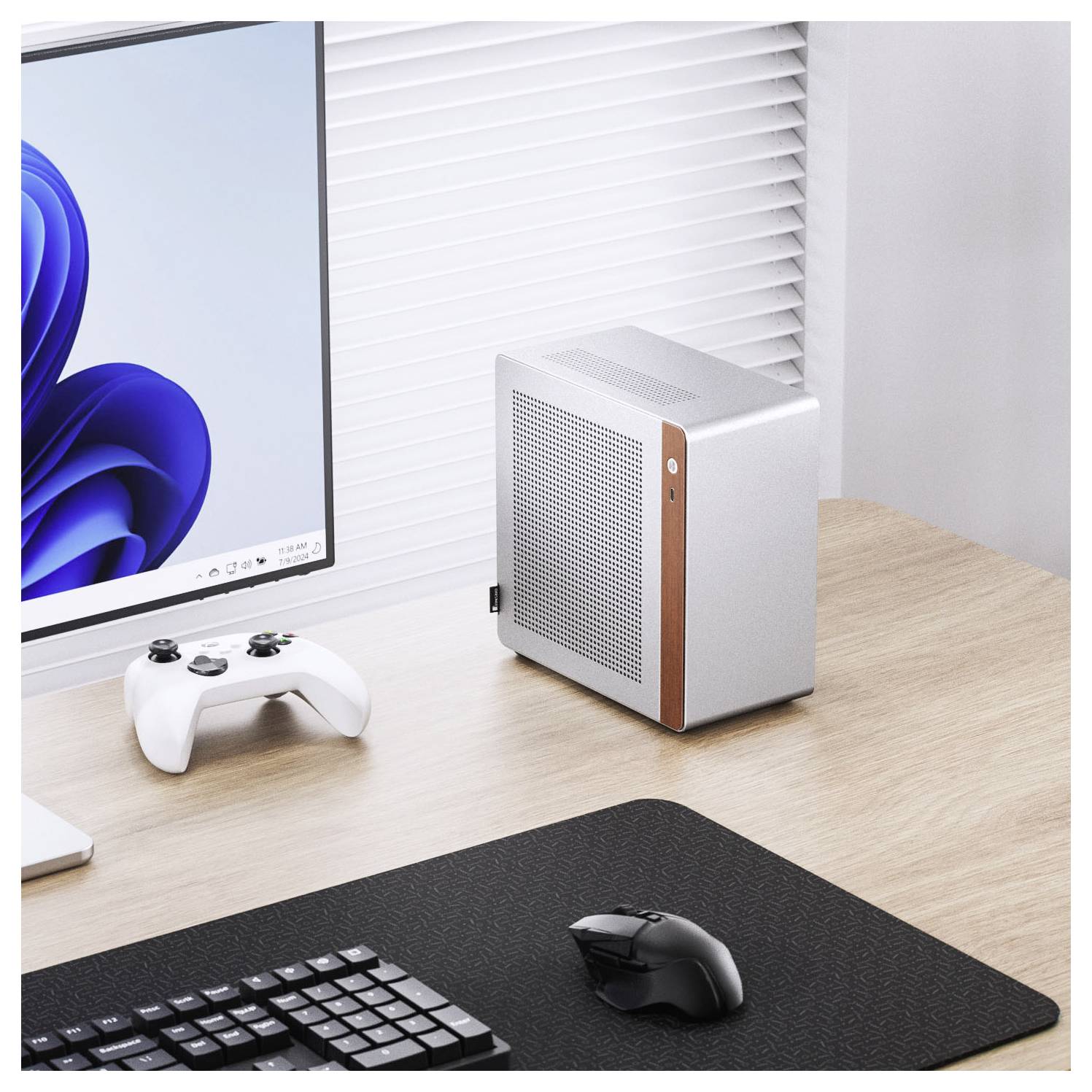 A compact, silver desktop computer next to a monitor displaying a blue swirl design. A white game controller and black keyboard are nearby.
