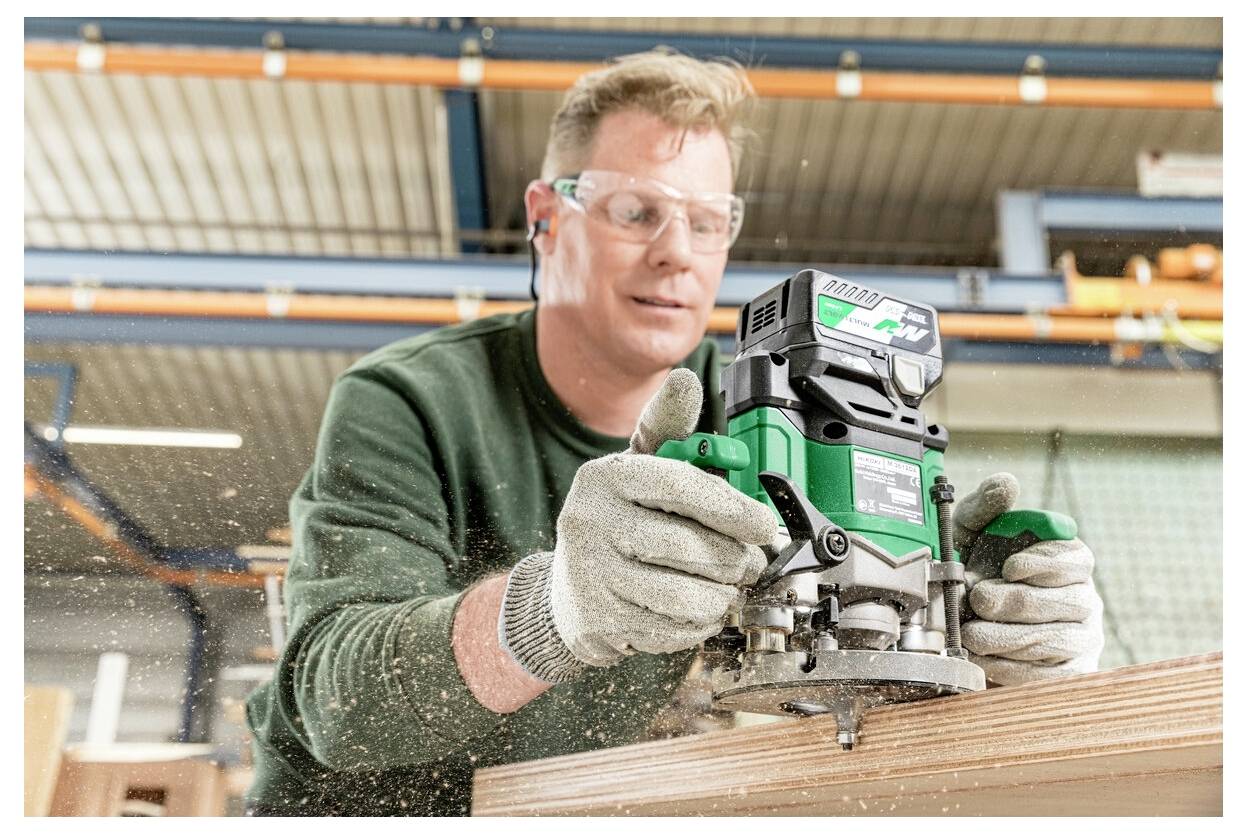 A person wearing protective eyewear uses a green power tool to shape the edge of a wooden plank in a workshop.