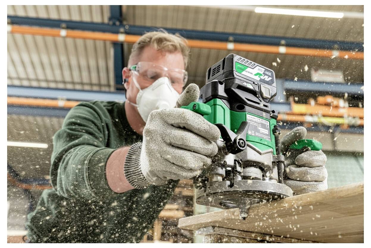 A person wearing safety gear operates a power tool on wood, creating a cloud of sawdust in an industrial setting.