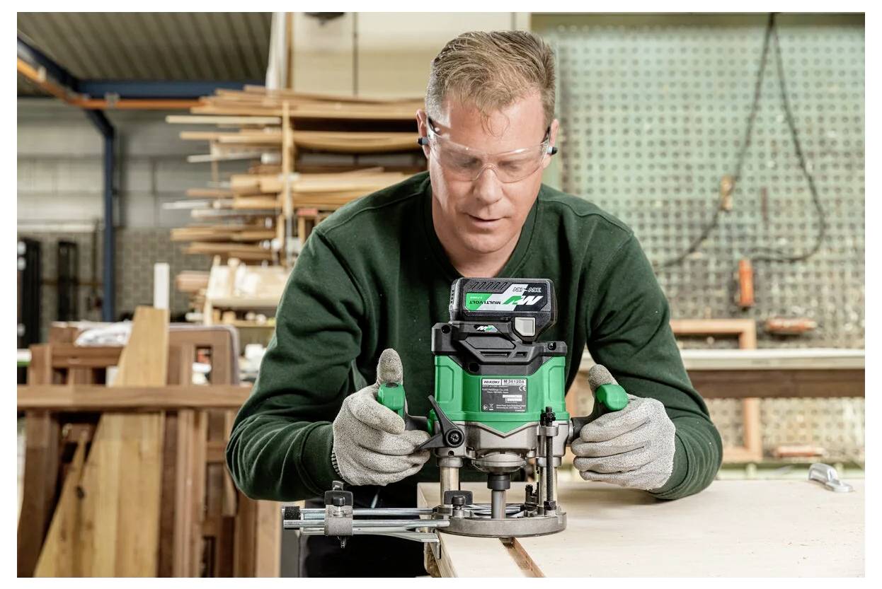 A person wearing safety gear uses a green power tool to shape wood in a workshop, surrounded by wooden planks and tools.