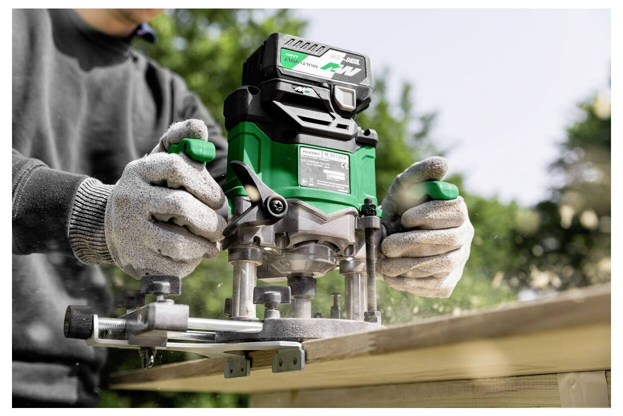 Person using a green power tool with a dust port, wearing gloves, working on a wooden board outdoors.