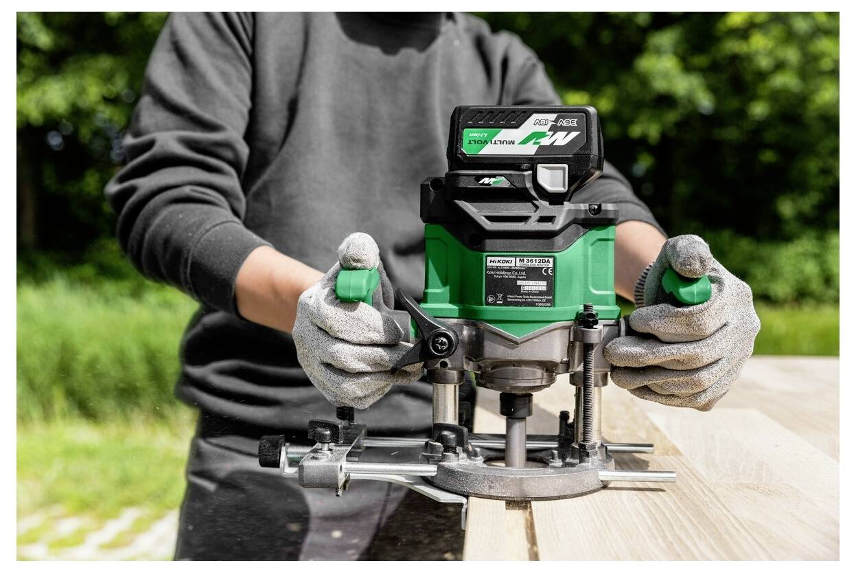 A person uses a green power tool to carve into a wooden surface outside, wearing gloves for safety.