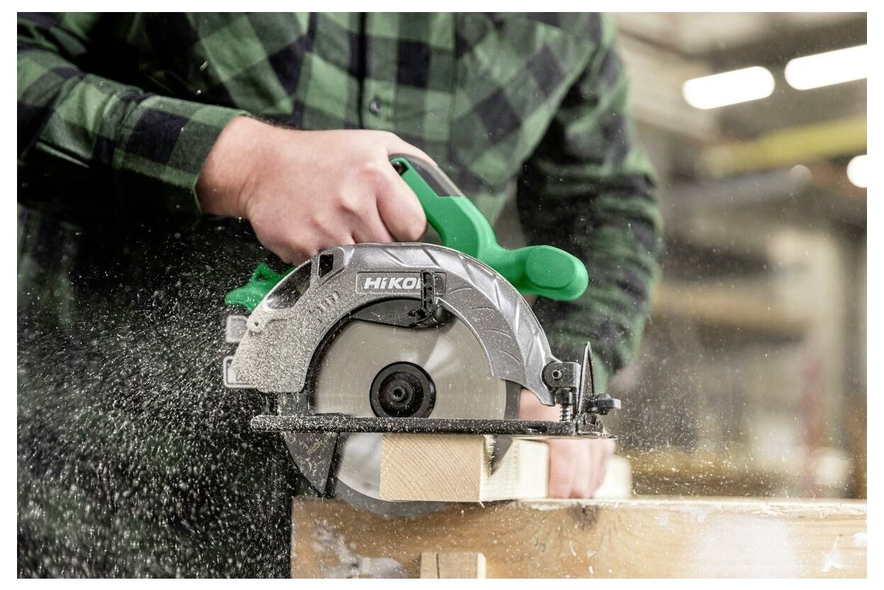 A person uses a circular saw to cut wood, creating dust. The green-handled saw is in motion, with sawdust spraying outward.
