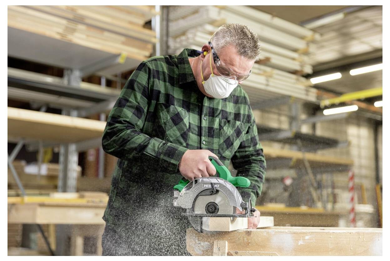 A person wearing safety gear uses a circular saw to cut a piece of wood in a workshop, surrounded by shelves and tools.