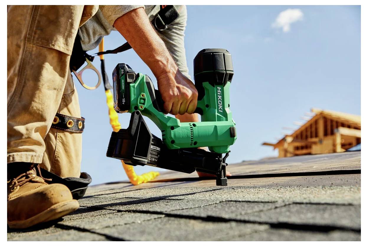 A person on a roof uses a green nail gun to secure shingles, with a wooden house frame in the background under a clear sky.