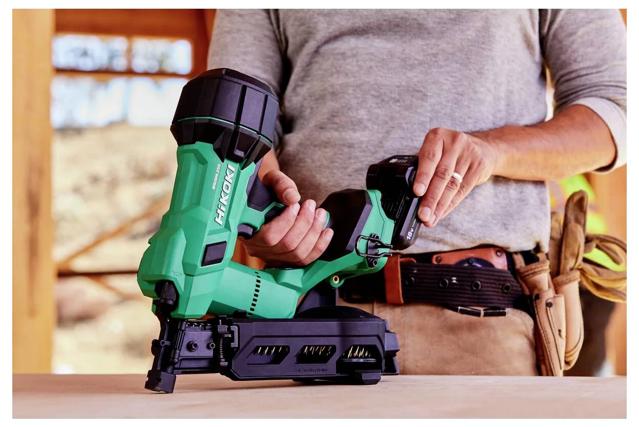 A person adjusts a green nail gun while standing at a workbench, wearing a tool belt, suggesting construction or woodworking work.