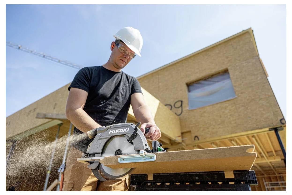 A construction worker wearing a hard hat and goggles uses a circular saw to cut wood in front of a partially built wooden house.
