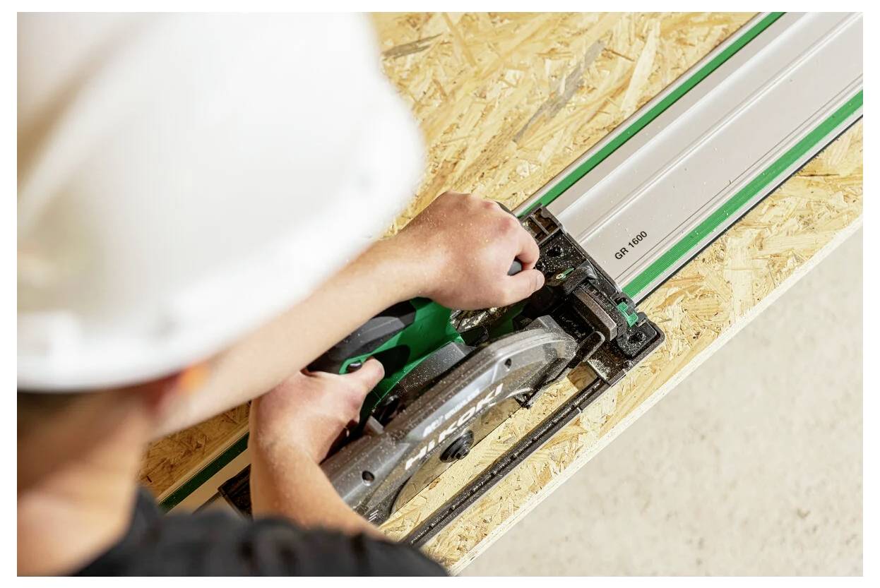 A worker in a white hard hat uses a circular saw on a wooden board. The saw is attached to a guide rail, ensuring a straight cut.