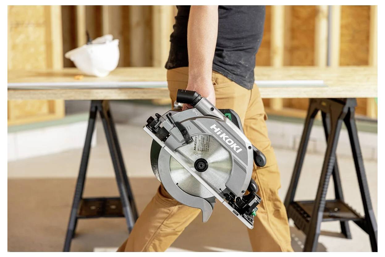 A person carries a Hitachi circular saw in a workshop, with wooden boards and saw horses visible. Construction context implied.