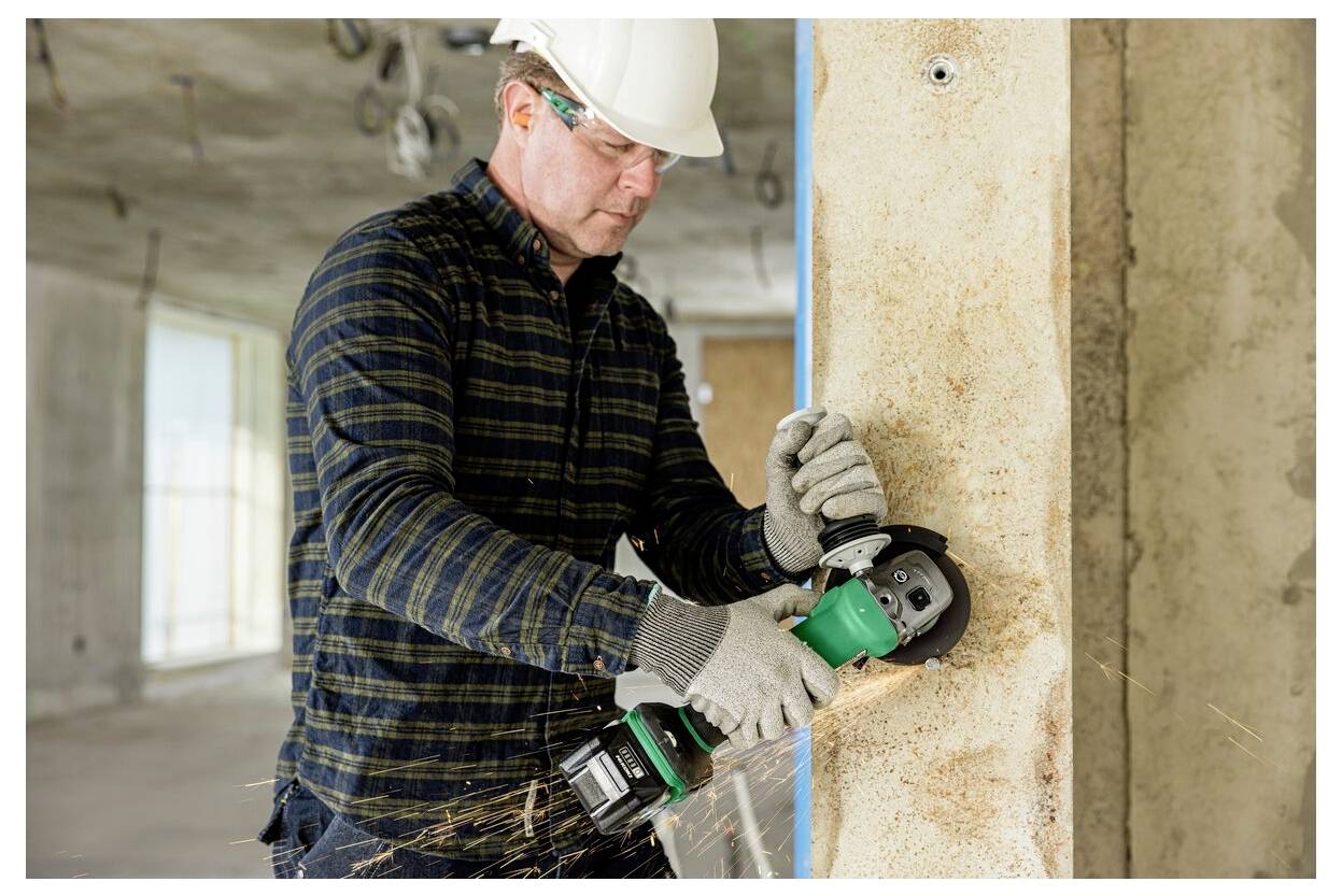 A construction worker wearing safety gear uses an angle grinder on a concrete wall, creating sparks in an unfinished building.