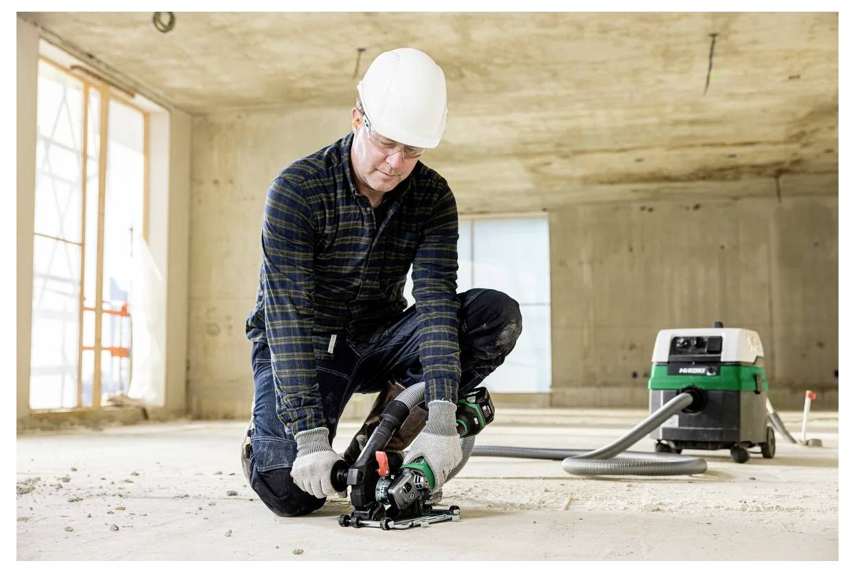 A construction worker, wearing a hard hat and gloves, uses a power tool on a concrete floor in a partially finished building.