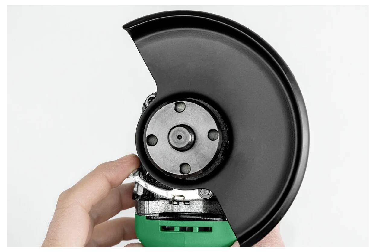 A hand adjusts the safety guard of a green angle grinder, focusing on the tool's circular blade area.