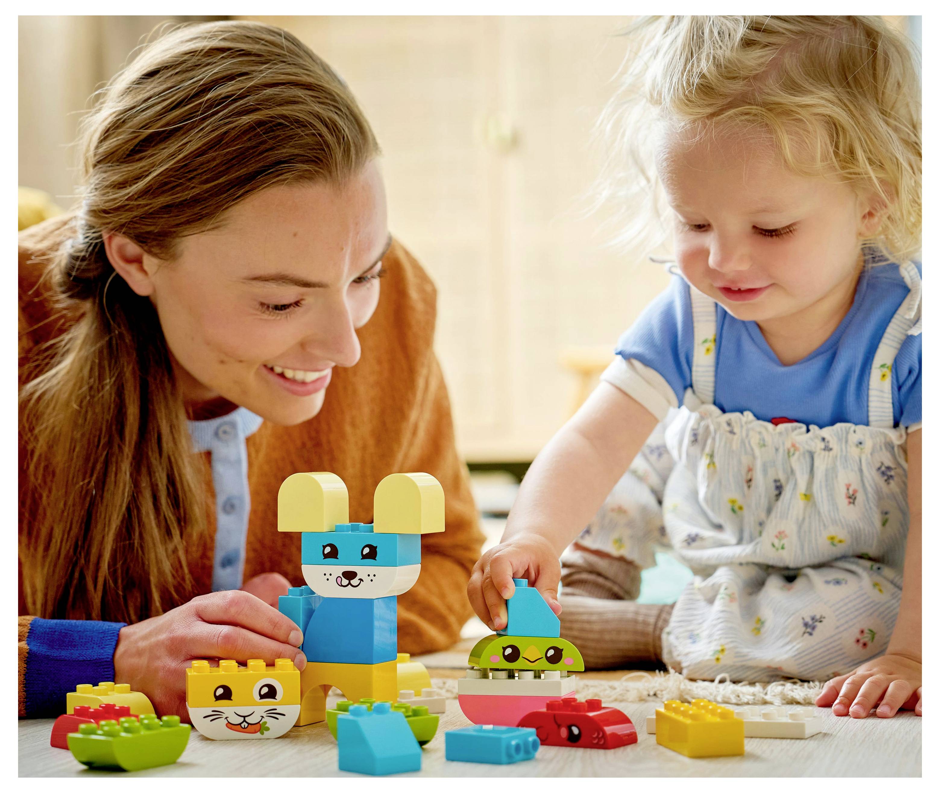 An adult and a child play with colorful building blocks on the floor. The child is smiling and focused on stacking the blocks.