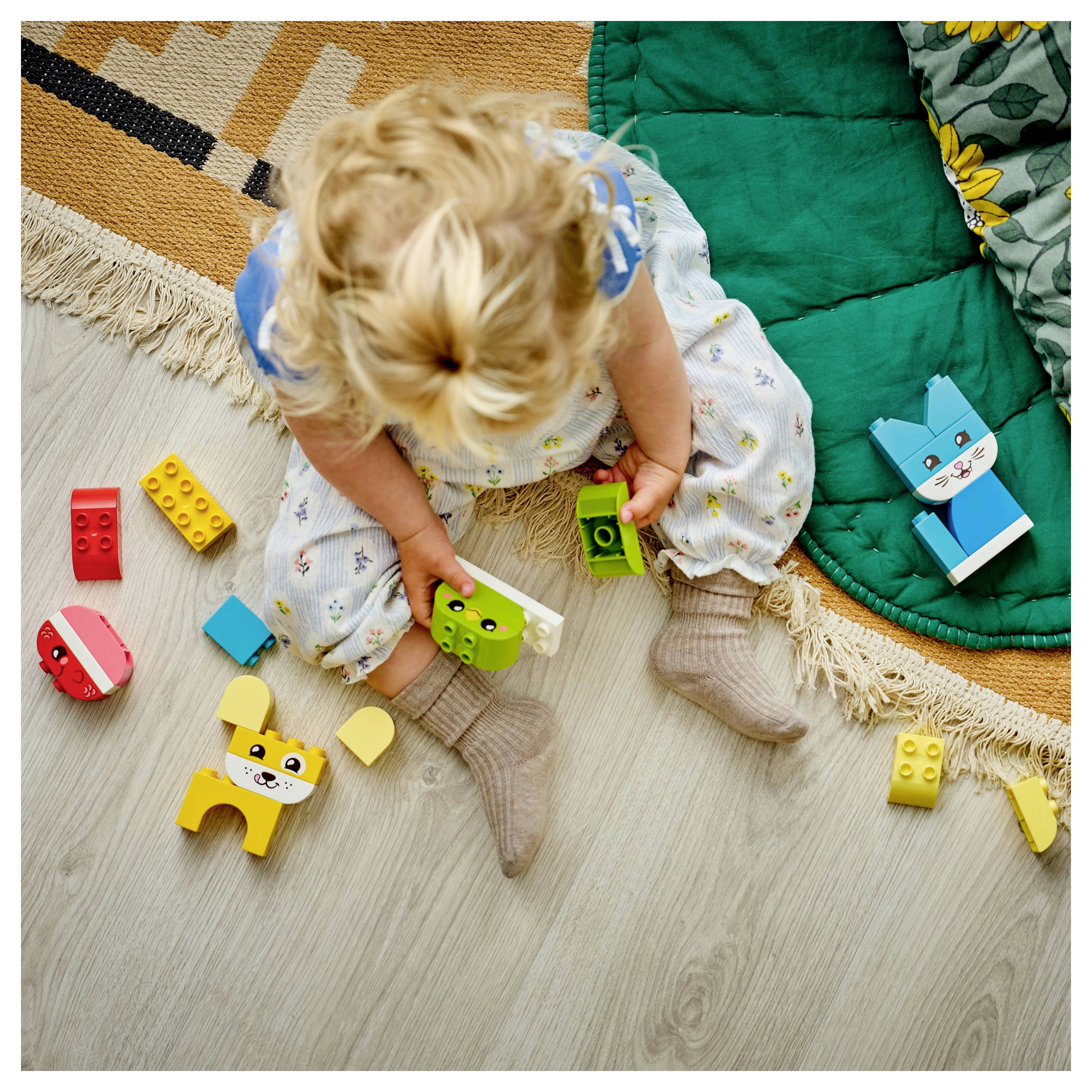 A toddler sits on the floor playing with colorful toy blocks shaped like animals, surrounded by a patterned rug and a green blanket edge.