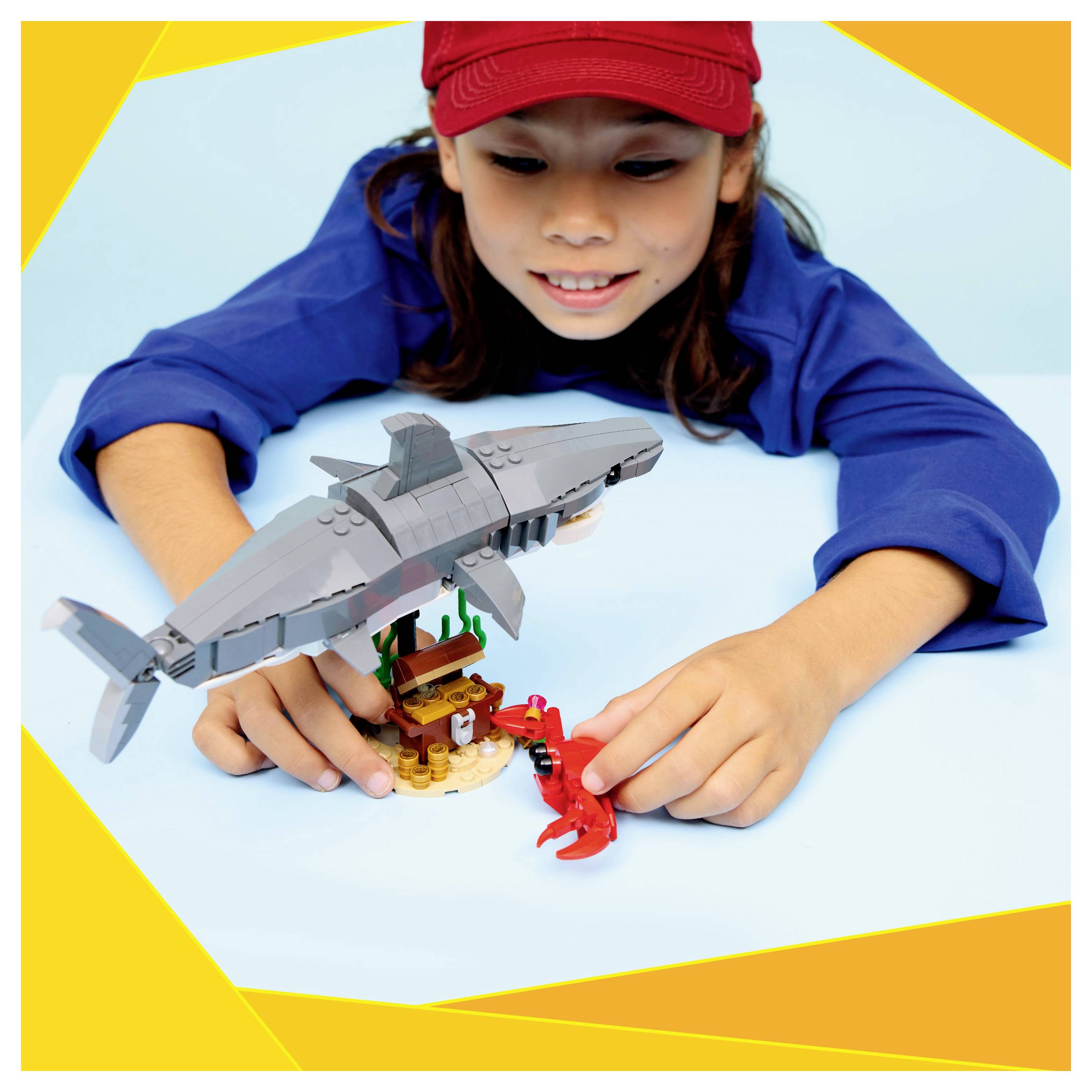 A child in a red cap and blue shirt plays with a toy set, featuring a model shark and sea-themed elements, on a light blue surface.