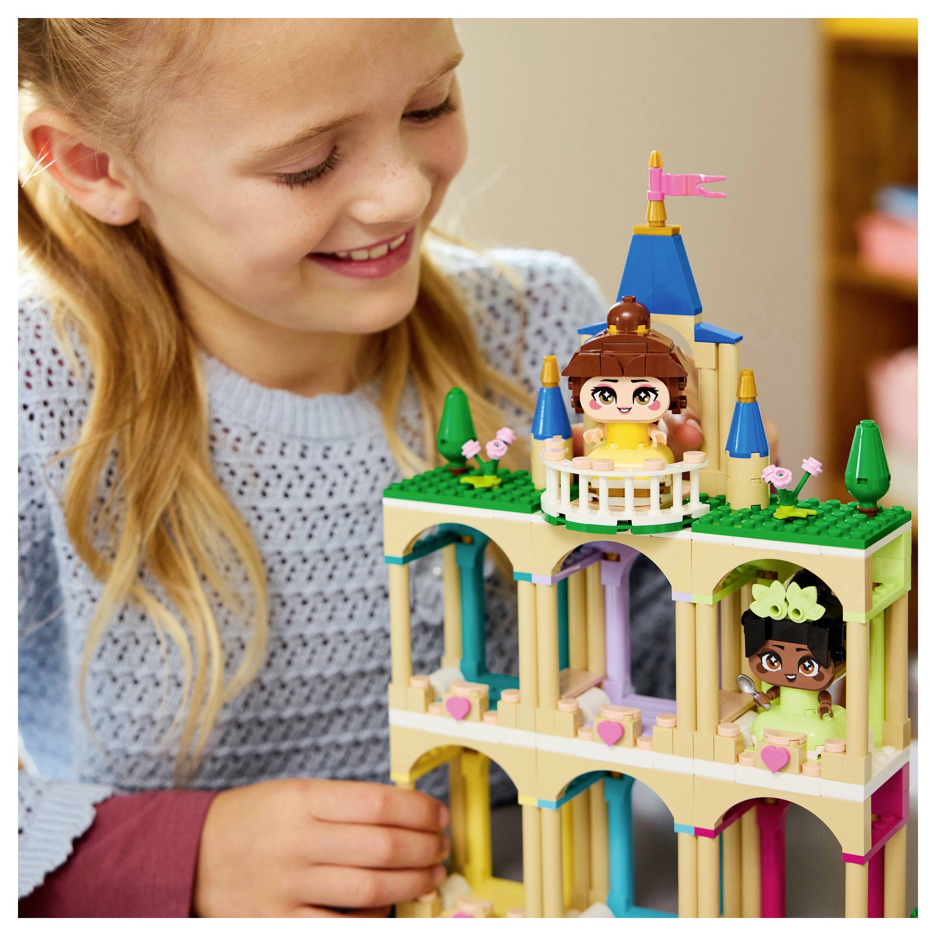 A child is smiling while playing with a colorful toy castle, featuring small toy figures and turrets, on a wooden table.