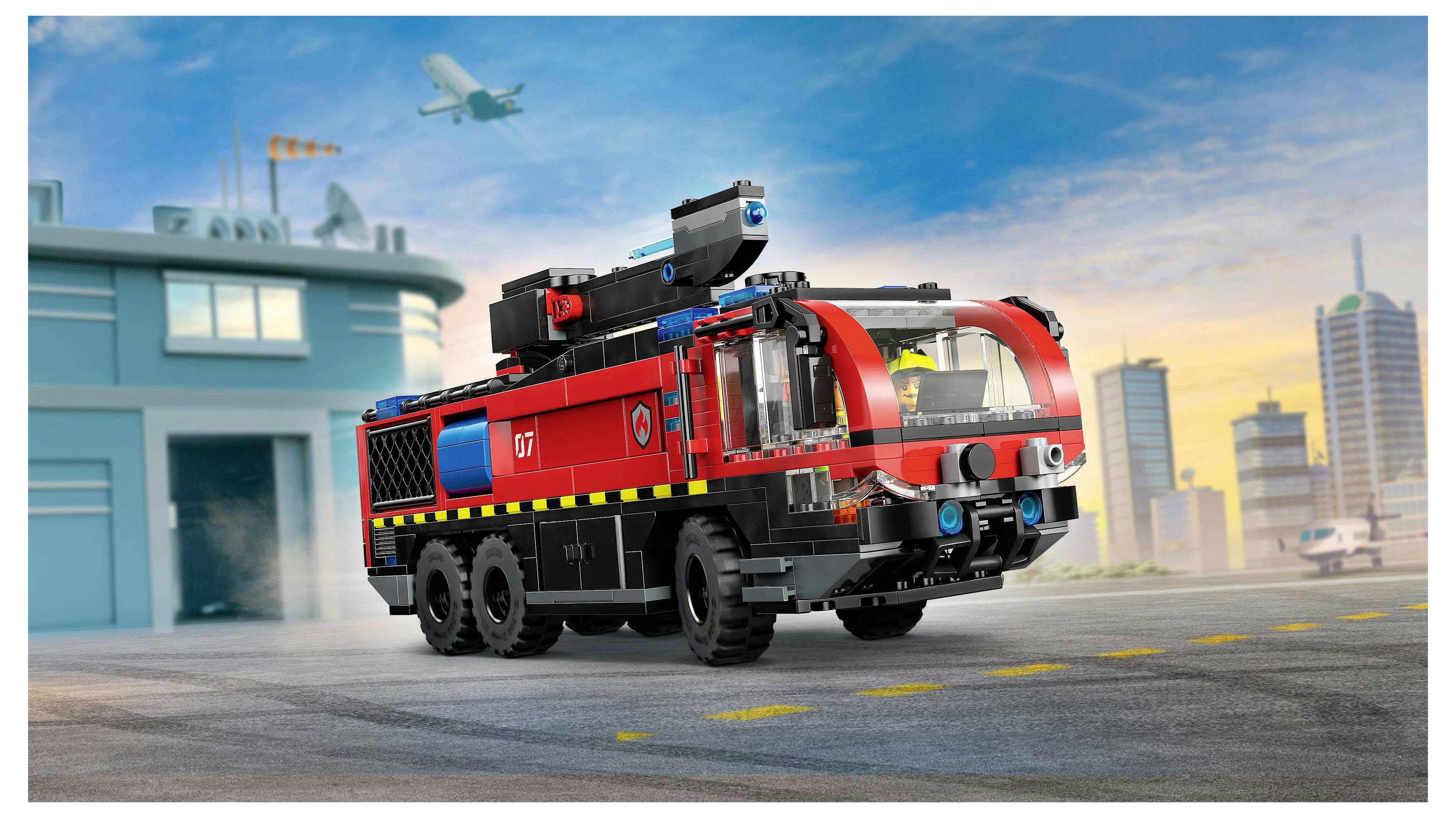 A toy fire truck on an airport runway with an airplane in the sky and buildings in the background, depicting a flying and city environment.