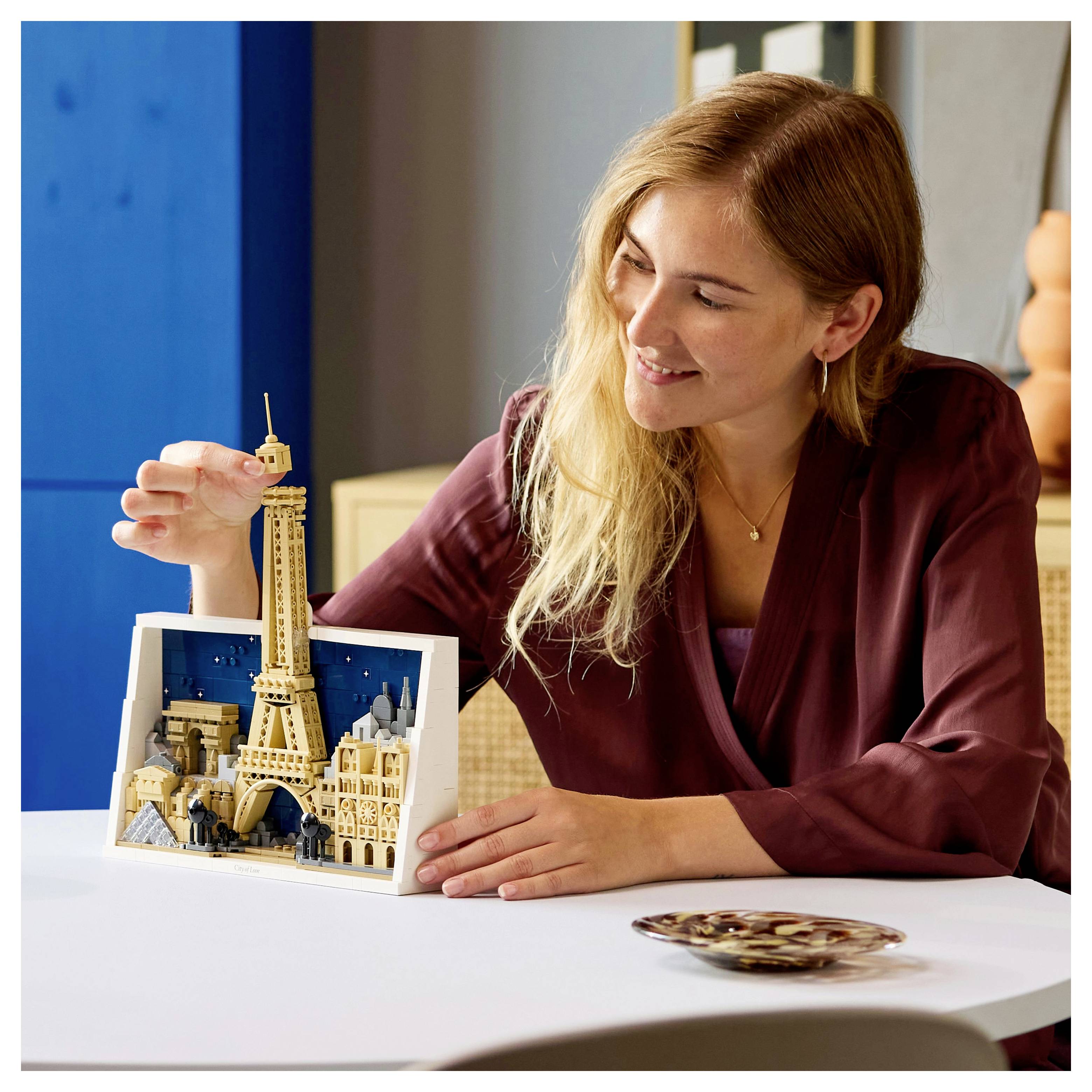 A person smiles while assembling a detailed miniature model of the Eiffel Tower on a table, indicating a creative and engaging activity.