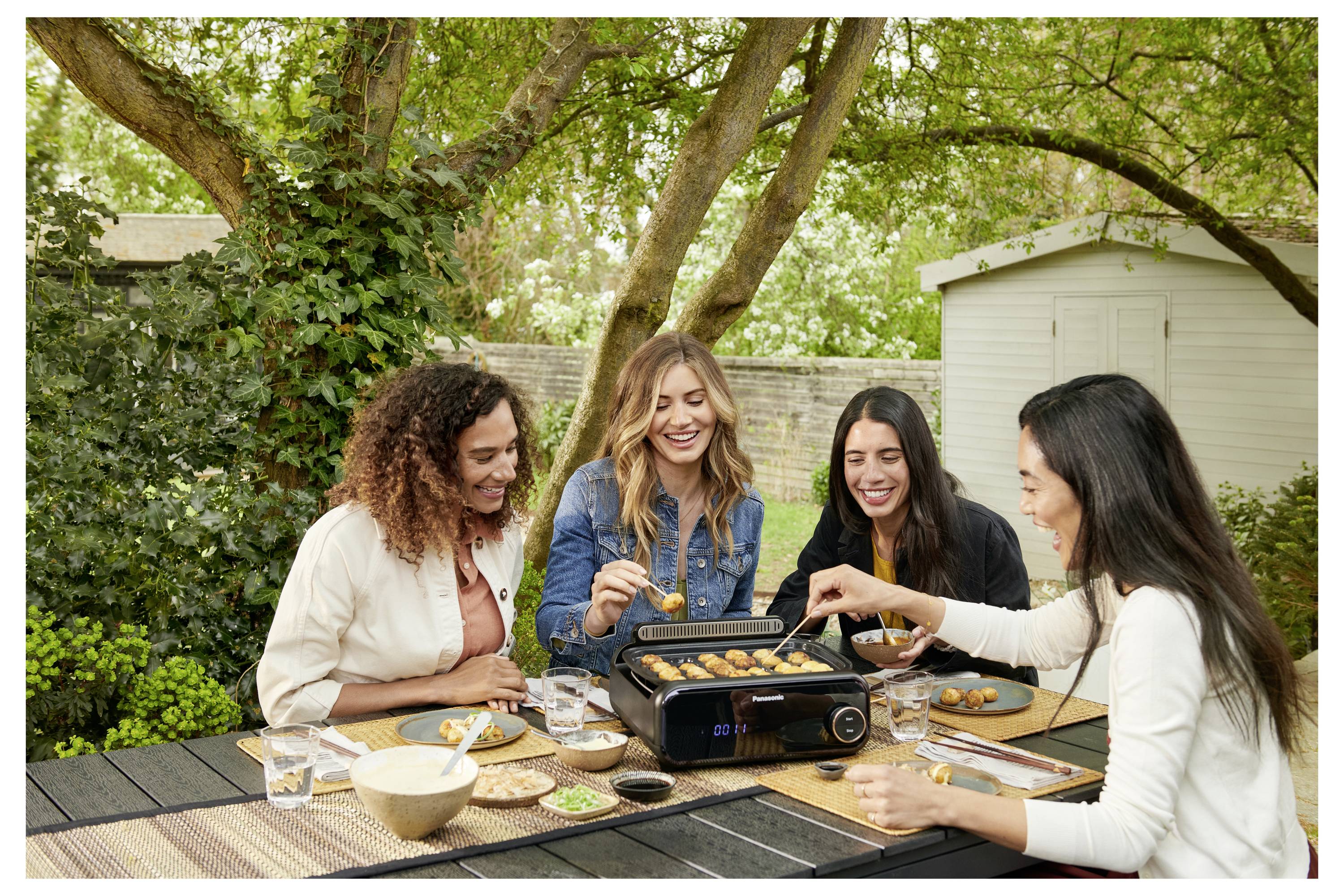 Four women enjoy cooking and eating at an outdoor table surrounded by greenery. A grill is placed on the table.