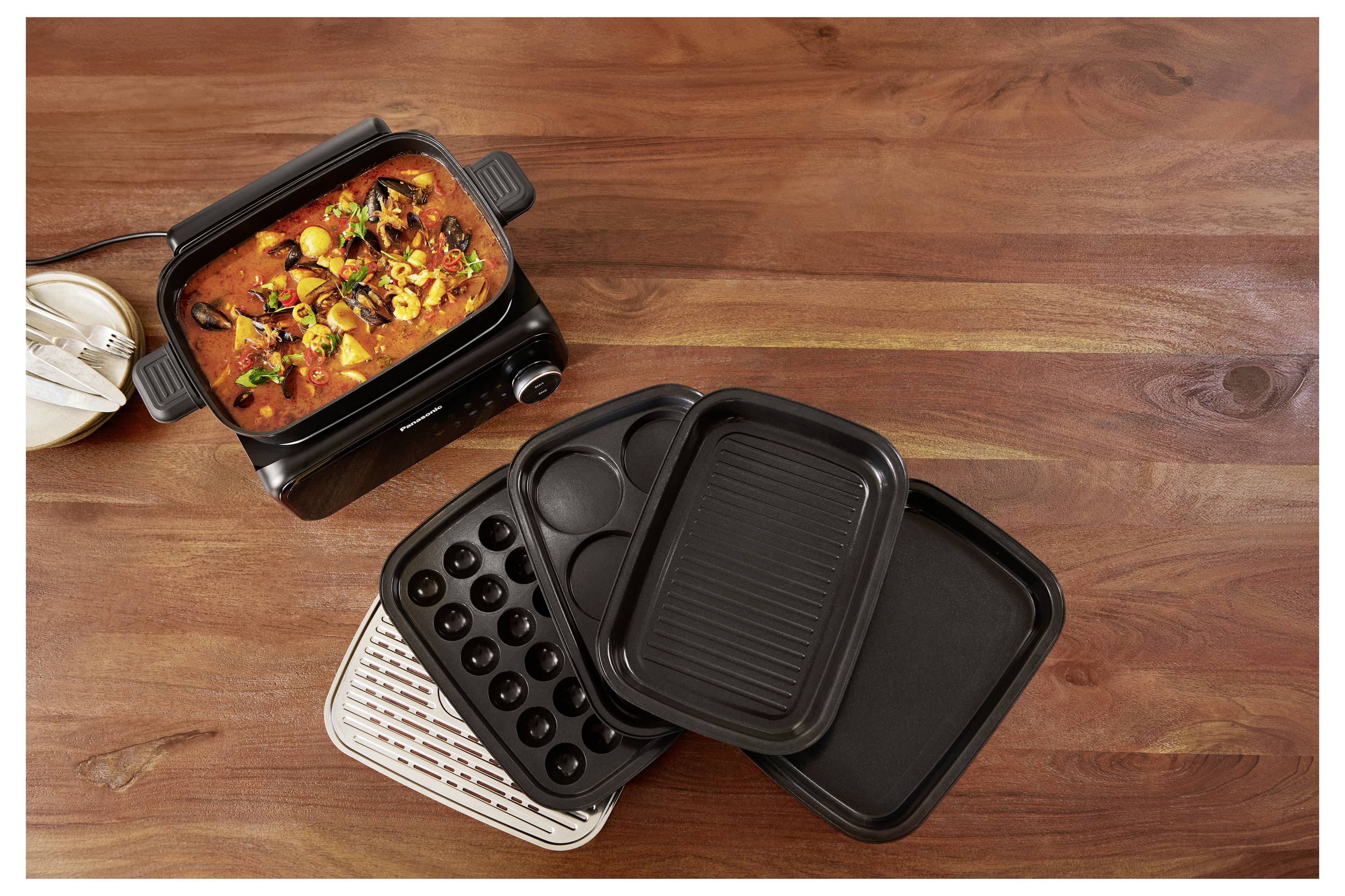 A multi-functional cooking appliance on a wooden surface, showing a pot with simmering stew, detachable grilling and baking trays nearby.