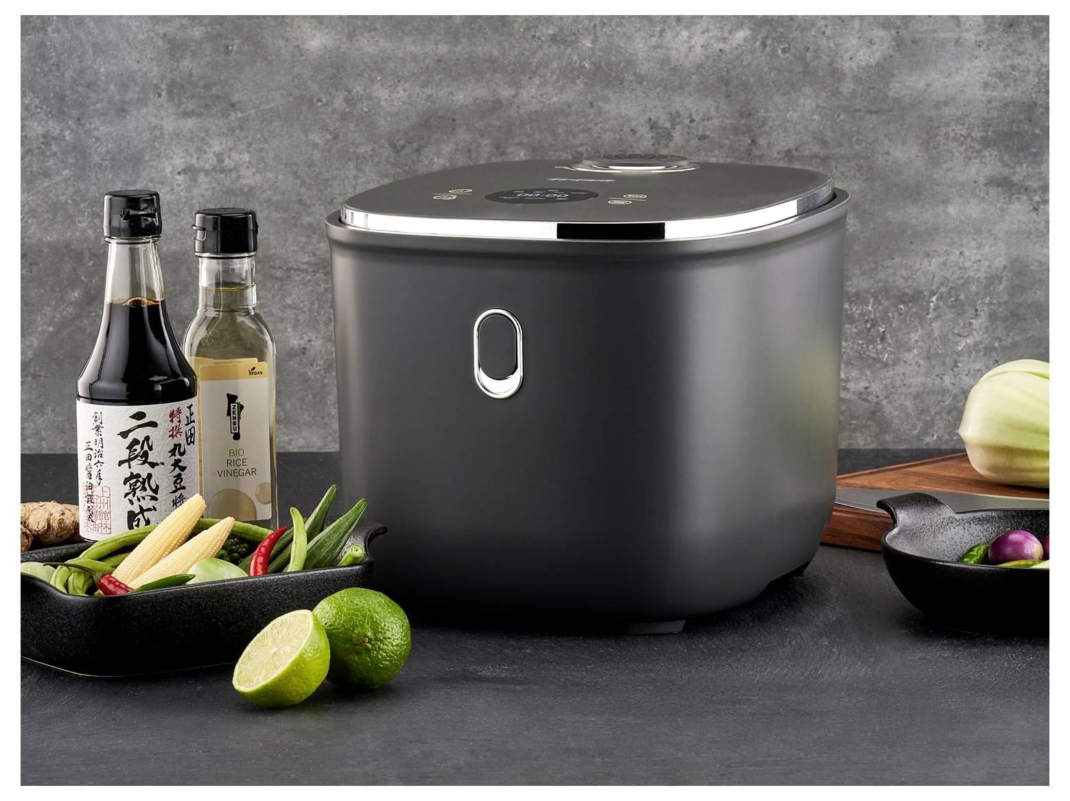 A black rice cooker on a kitchen counter with bottles of sauce, vinegar, and assorted vegetables, including lime and ginger, nearby.