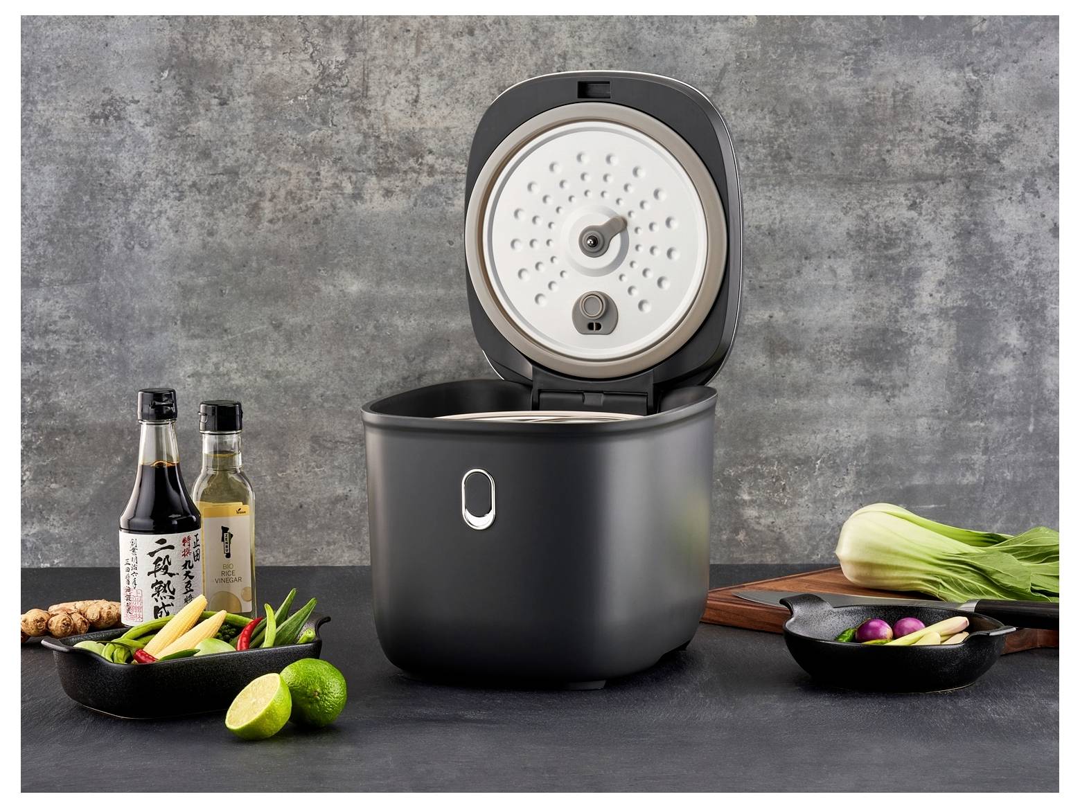 A black rice cooker with its lid open is centered on a kitchen counter, surrounded by fresh vegetables, sauces, and spices.