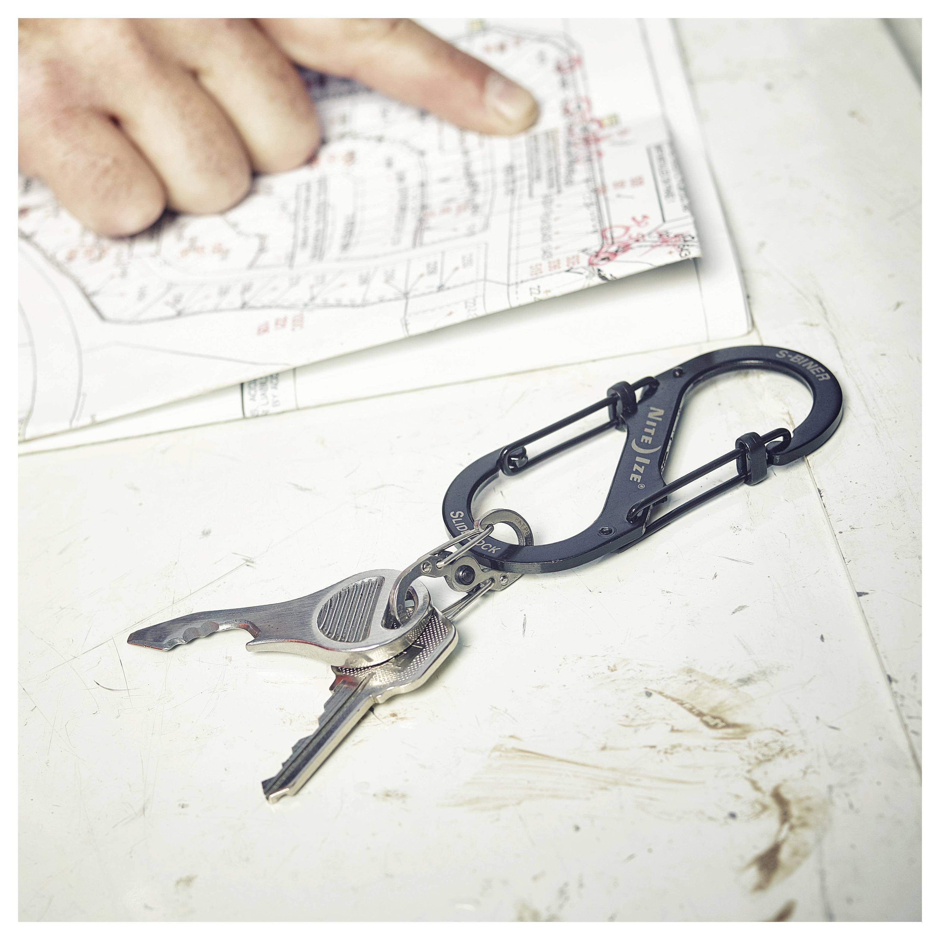 A hand points at a building plan on paper. Nearby, a black carabiner holds keys and a multitool, resting on a white surface.