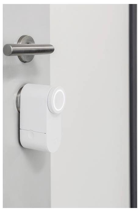 A white smart lock is installed on a modern door, featuring a cylindrical handle on the left. The lock has a sleek, minimalist design.