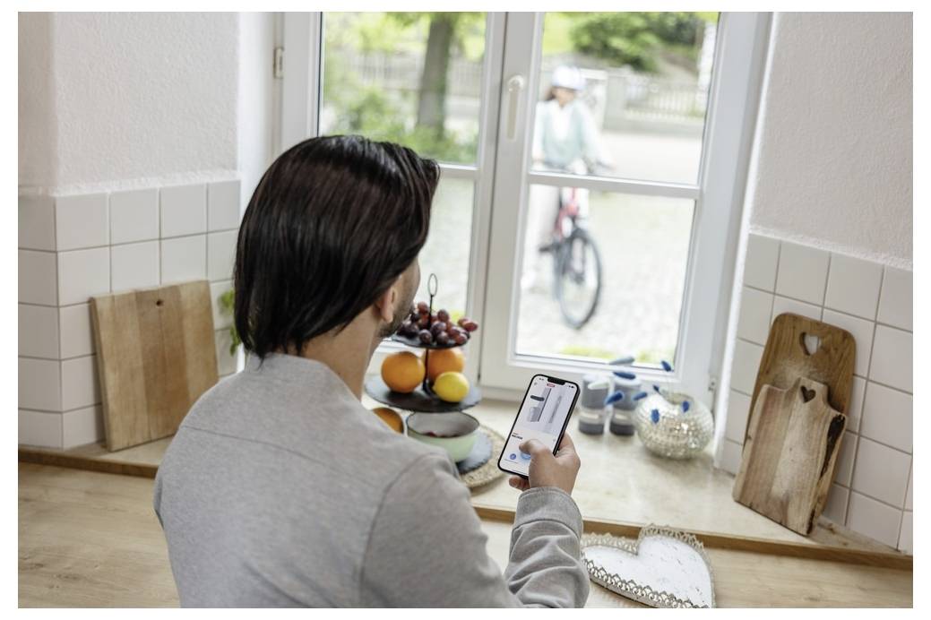 A person in a kitchen looks at a smartphone displaying a video feed. Through the window, a child on a bicycle is visible outside.