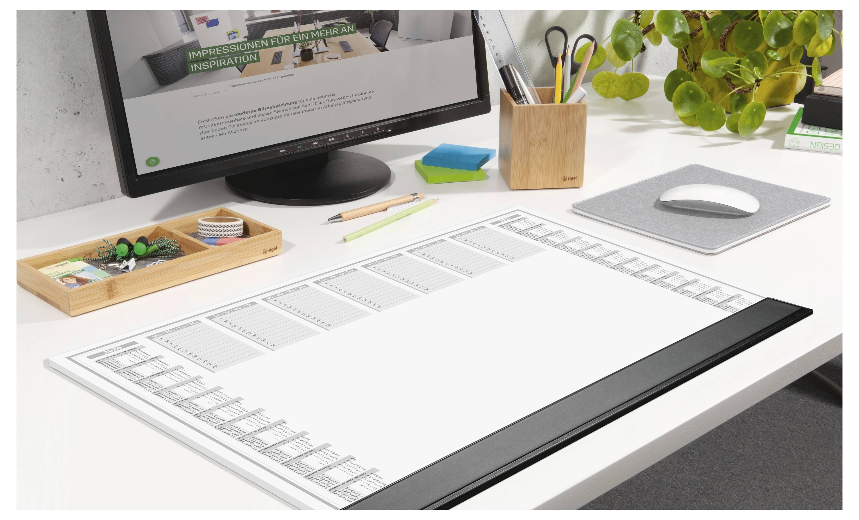 A clean, organized desk with a computer, calendar planner, stationery tray, and potted plant, suggesting a workspace for planning or creativity.
