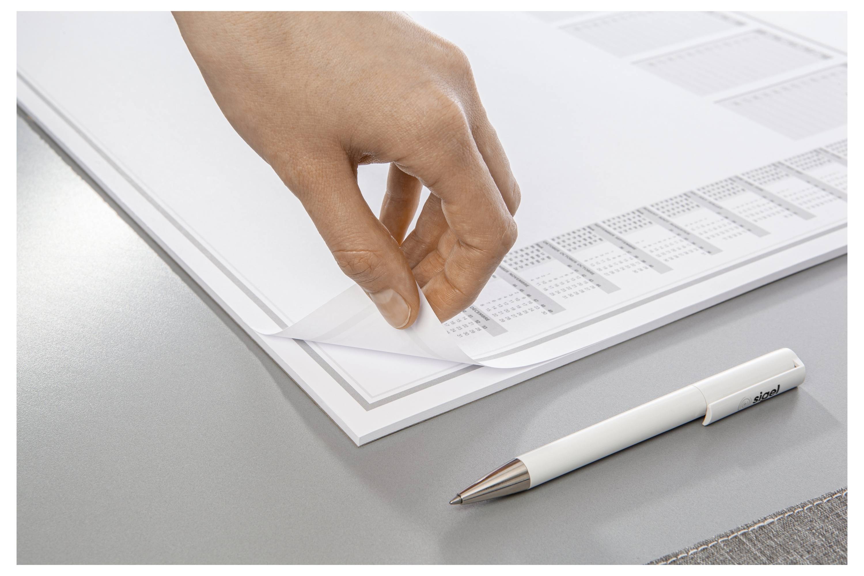 A hand lifts the corner of a stack of white papers on a desk next to a white and silver pen, suggesting organization or note-taking.