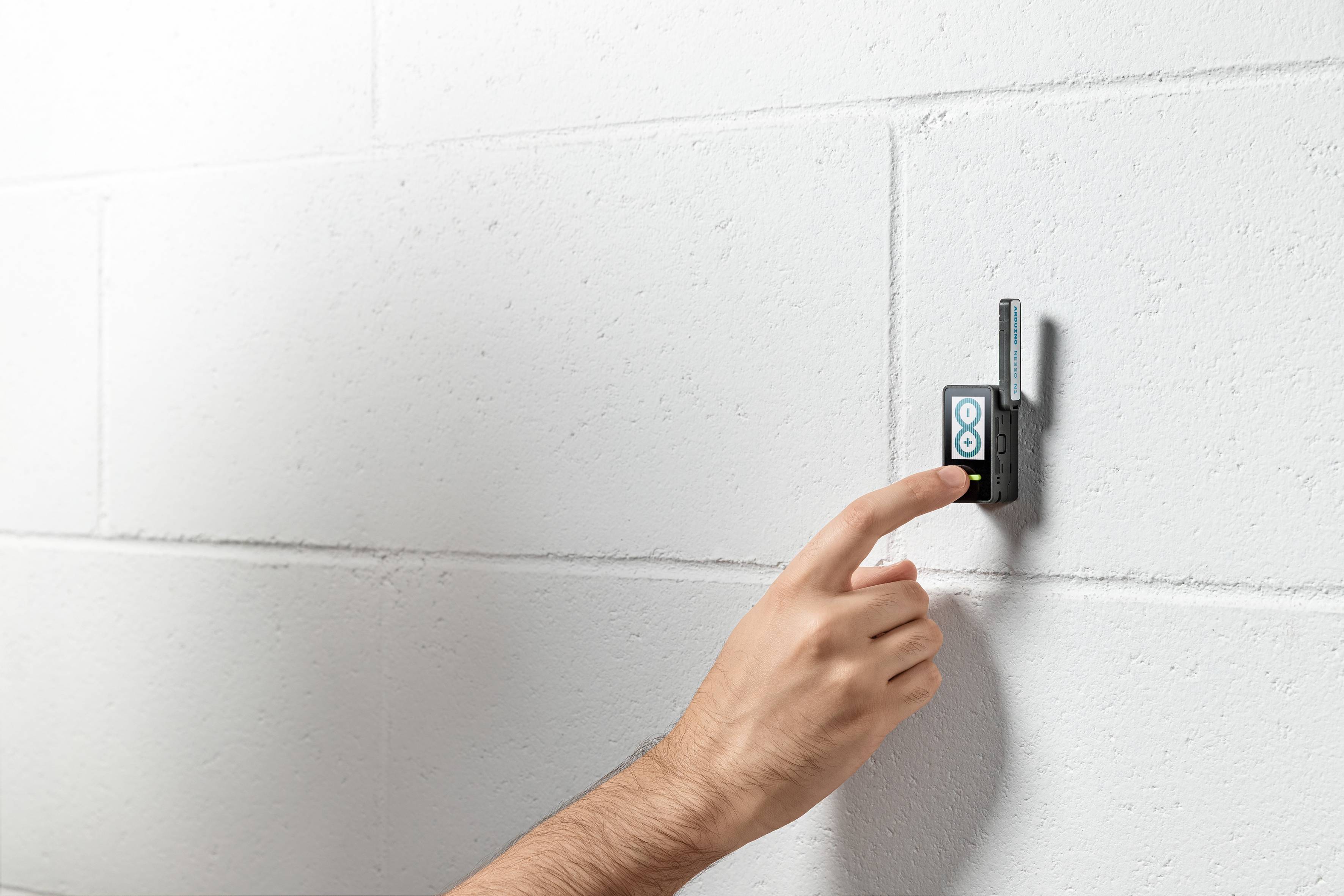 A hand presses a button on a small black device with an antenna, mounted on a white brick wall.