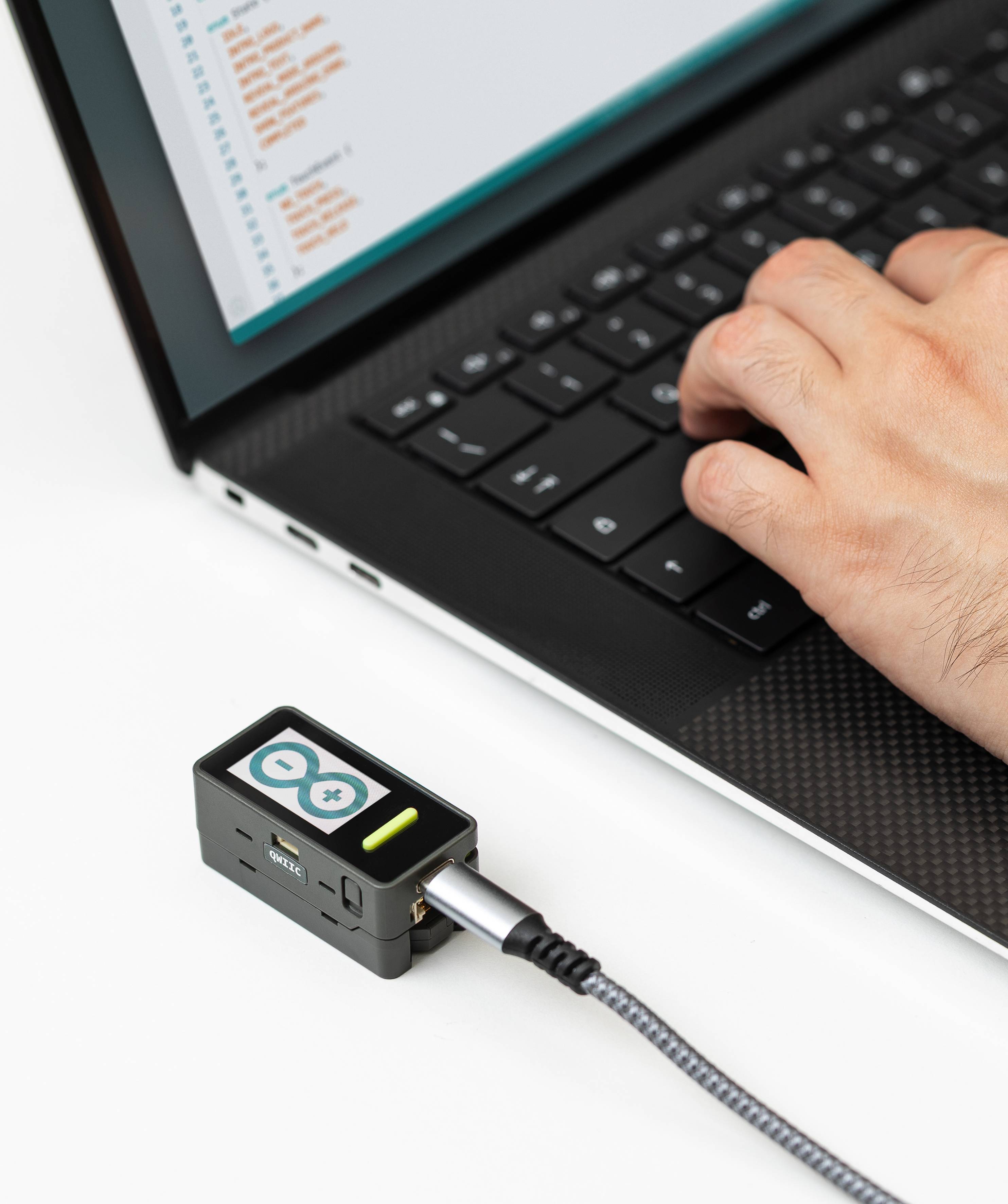 Person typing on a laptop with coding software on the screen, connected to an Arduino board via a USB cable.