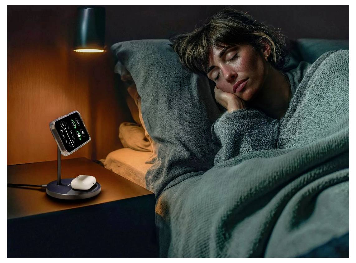 A woman sleeps in bed under a green blanket. A mobile phone on a bedside stand shows health monitoring data under a small lamp.