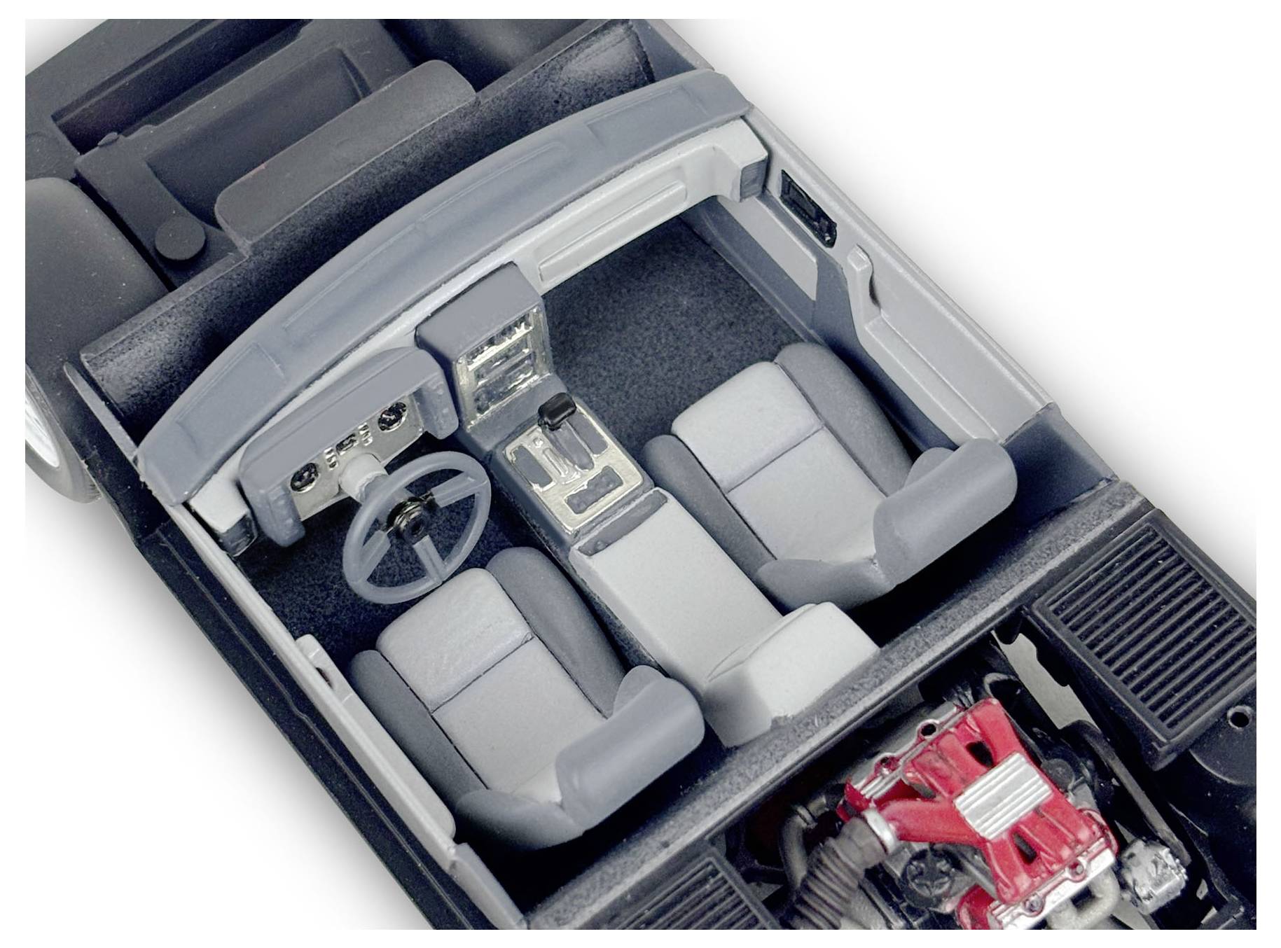 Model car interior showing two gray seats, a steering wheel, and center controls, with a visible engine detail in red at the back.