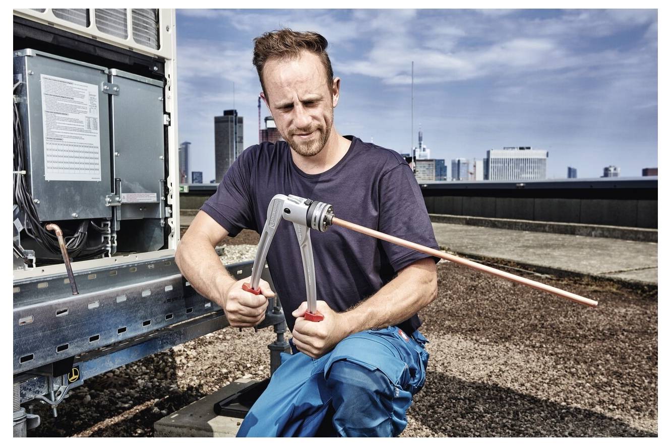 A person uses a specialized tool on a rooftop near industrial equipment, with a city skyline in the background.