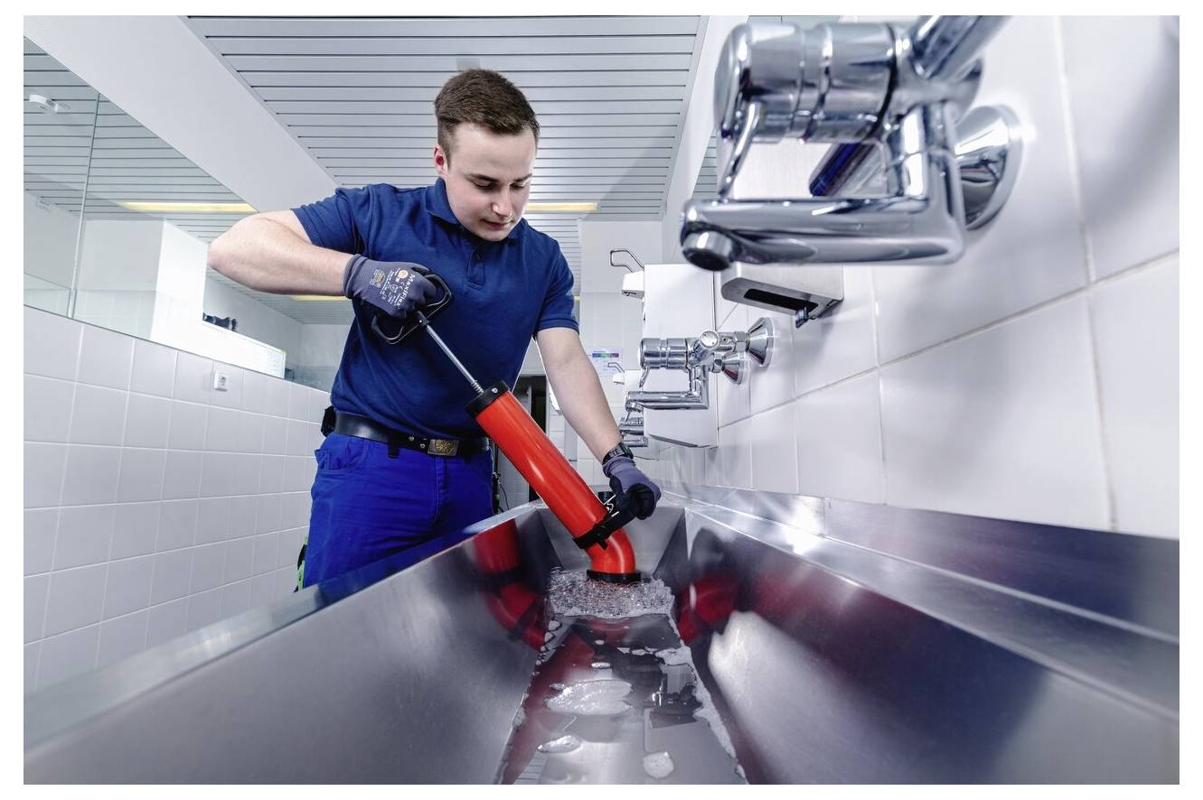 A plumber in a blue uniform uses a plunger to clear a long, stainless steel sink clogged with water in a commercial restroom.