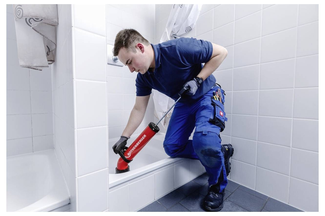 A plumber in blue overalls uses a silicone gun to seal the edge of a bathtub in a tiled bathroom, focusing on precision.