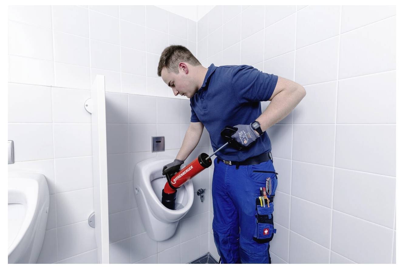 A plumber in uniform uses a tool to unclog a wall-mounted urinal in a tiled restroom. He wears gloves and has tools in his pocket.
