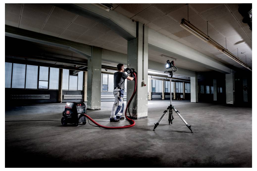 A person in work attire uses an industrial vacuum in a large, empty, industrial space, with a tripod-mounted light aimed at the work area.
